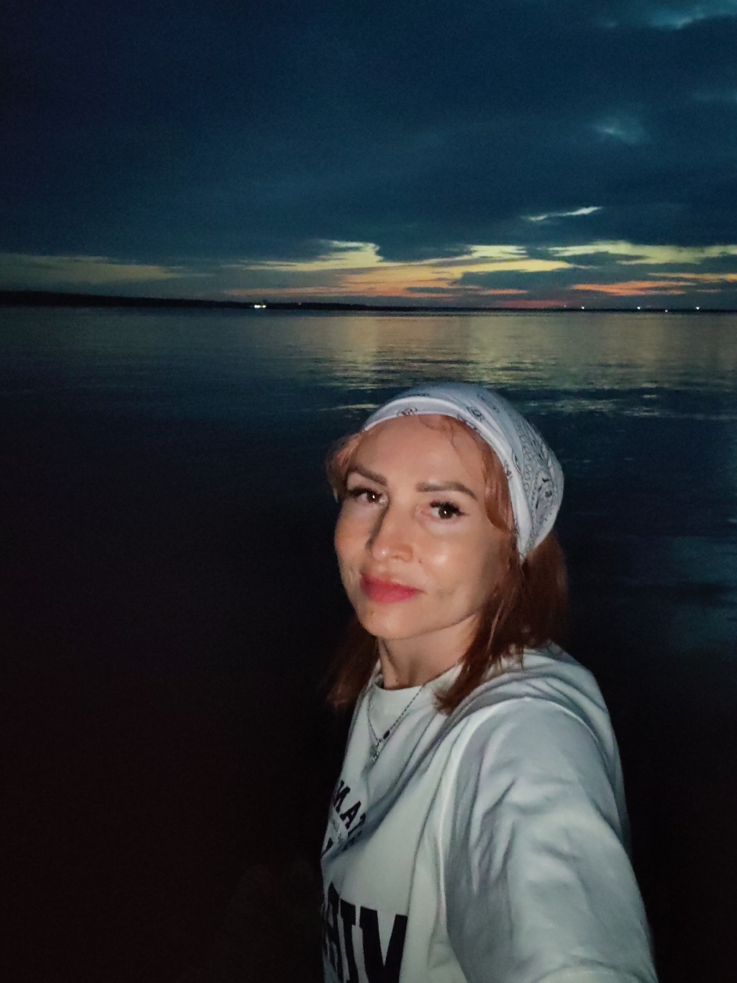 A selfie of a young woman with auburn hair and a white bandana is taken at dusk by a body of water. She is wearing a white sweatshirt with text on it. The sky is dark blue with streaks of orange and yellow from the setting sun. The water reflects the sky and appears calm. The location is likely the Kama River in Chaikovsky, Russia, known for its scenic riverbanks and opportunities for recreation.