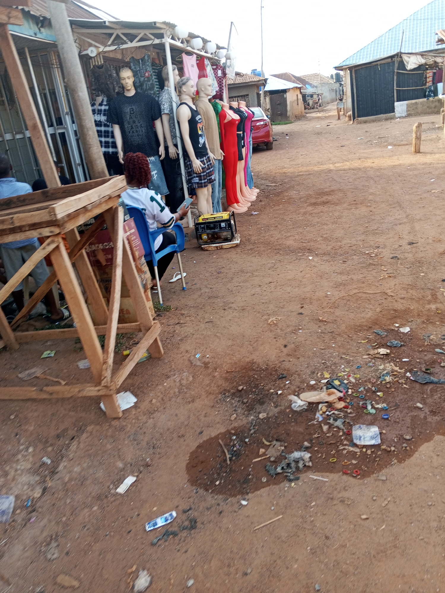 The image shows a street scene in what appears to be a market or commercial area, possibly in Abuja, Nigeria.
**Setting:** A dirt road with visible litter. There are several buildings and shops lining the road.
**People:** Several people are present. A woman is seated in a blue chair looking at her phone. Two men are seated nearby. Another person is walking further down the road.
**Objects:** There are mannequins dressed in various clothing items. A portable generator is on the ground. A wooden structure is in the foreground. Trash, including plastic waste, is scattered on the ground.
**Action:** The woman is using her phone. People are present in the market or commercial area.
**Environment:** The overall environment suggests a developing area, with the presence of litter indicating a possible lack of waste management infrastructure.