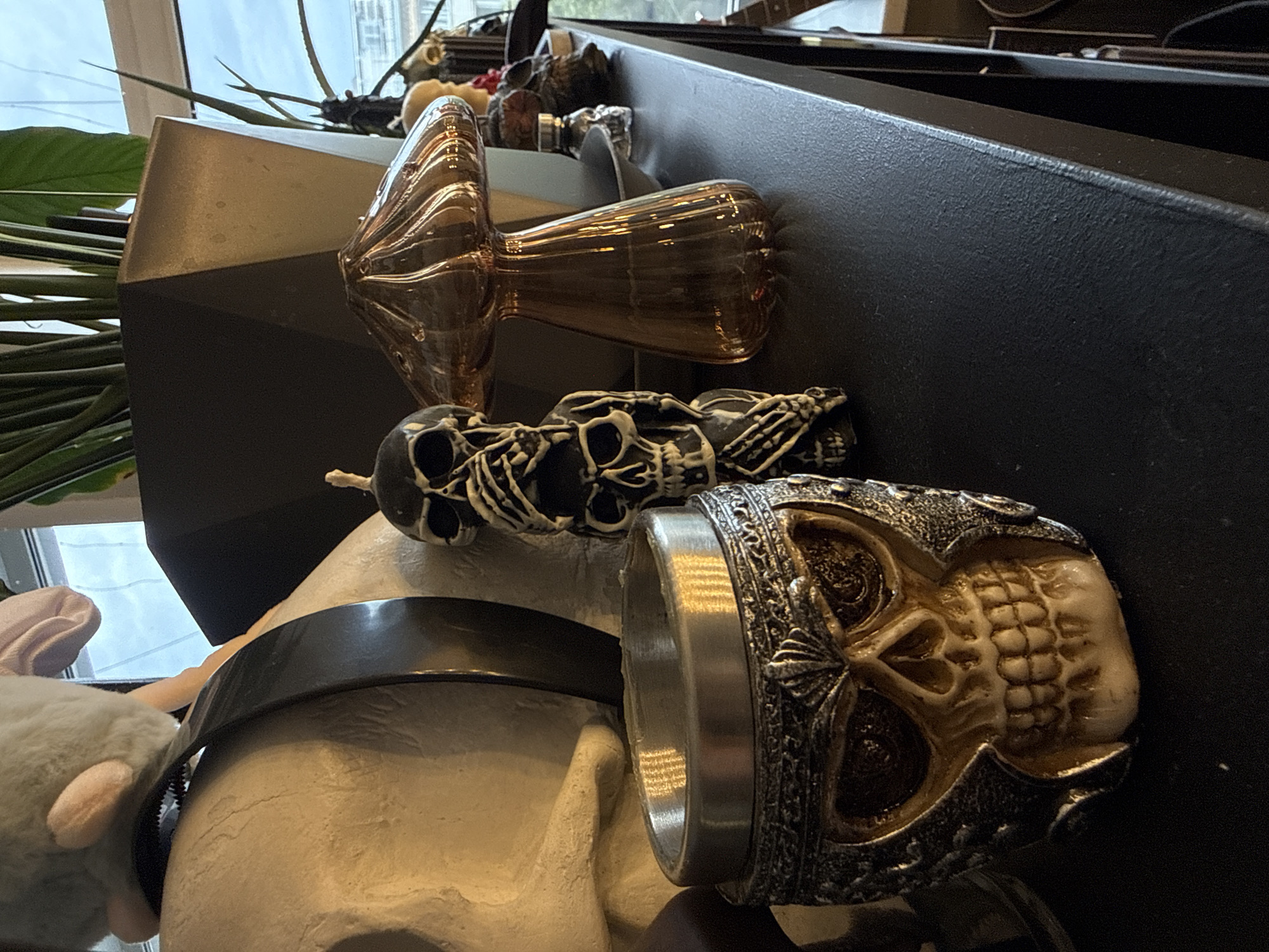 A still life arrangement featuring decorative skulls and an Edison-style light bulb rests on a surface, possibly a shelf. The skulls exhibit varying designs and colors, with one prominently displaying metallic accents. A mannequin head with a black band is partially visible. A plush toy with pink ears is present near the head. The background includes a window and indistinct urban elements, suggesting an indoor setting. The light suggests the photograph was taken in Krasnoyarsk, Russia.