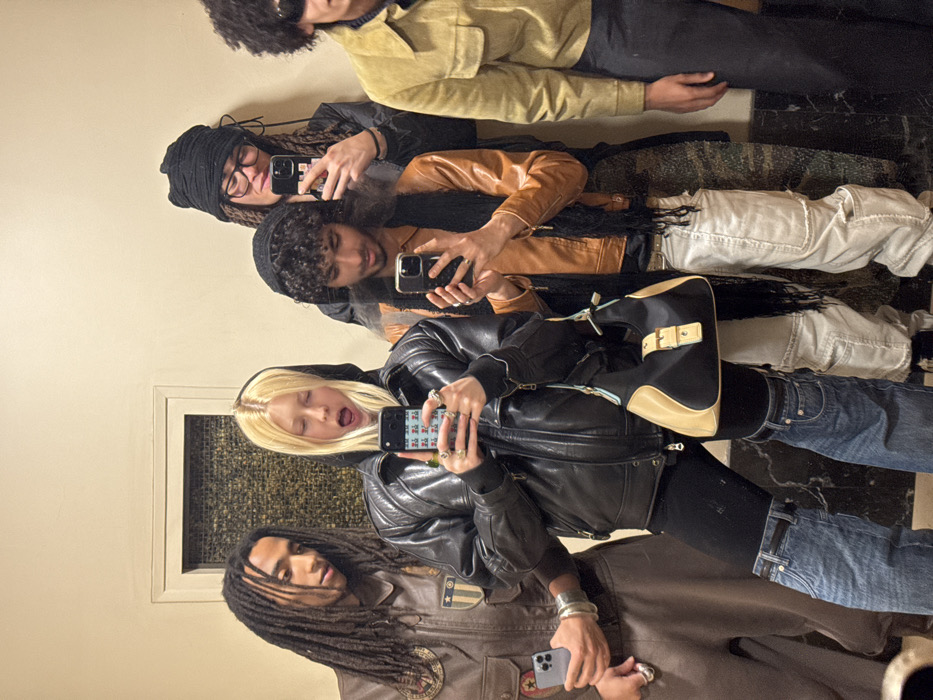 Five individuals are depicted indoors, taking a group mirror selfie in an unspecified location within Paris, France. The environment features light-colored walls, a framed recessed panel with dark textured material, and polished light-colored flooring.
From left to right:
1. A male with long dark dreadlocks, wearing a brown jumpsuit with patches on the chest (one resembling an Italian flag shield and another a star and stripes), stands looking down to his right. He wears a prominent silver cuff on his left wrist and holds a grey smartphone with three camera lenses in his left hand, its screen illuminated.
2. A female with blonde hair and dark roots, partially covered by a black hoodie, stands centrally with her mouth open. She wears a black leather bomber jacket over a black garment, and black leggings with blue jeans cut and worn over the leggings, secured with belts around her upper thighs. She has multiple rings on her fingers and holds a black smartphone with a patterned case in both hands, aiming it towards the mirror. A black and beige shoulder bag with a gold buckle hangs from her right arm.
3. A male with dark curly hair and a black knit beanie, wearing a tan leather jacket, a black shirt, and light-colored cargo pants, stands with a long black fringed scarf around his neck. He holds a black smartphone in his left hand, also aiming it towards the mirror. He also wears rings on his fingers.
4. Partially obscured behind the second male, another male with long dark dreadlocks or braids, wearing a dark knit cap/headwrap and glasses, holds a black smartphone in his right hand, aiming it towards the mirror.
5. On the far right, a partially visible male with dark curly hair and sunglasses is present, wearing a light olive green/yellow suede-like jacket and dark pants.
All individuals are positioned close together, actively engaged in the act of photographing themselves collectively using multiple smartphones directed towards the reflective surface.