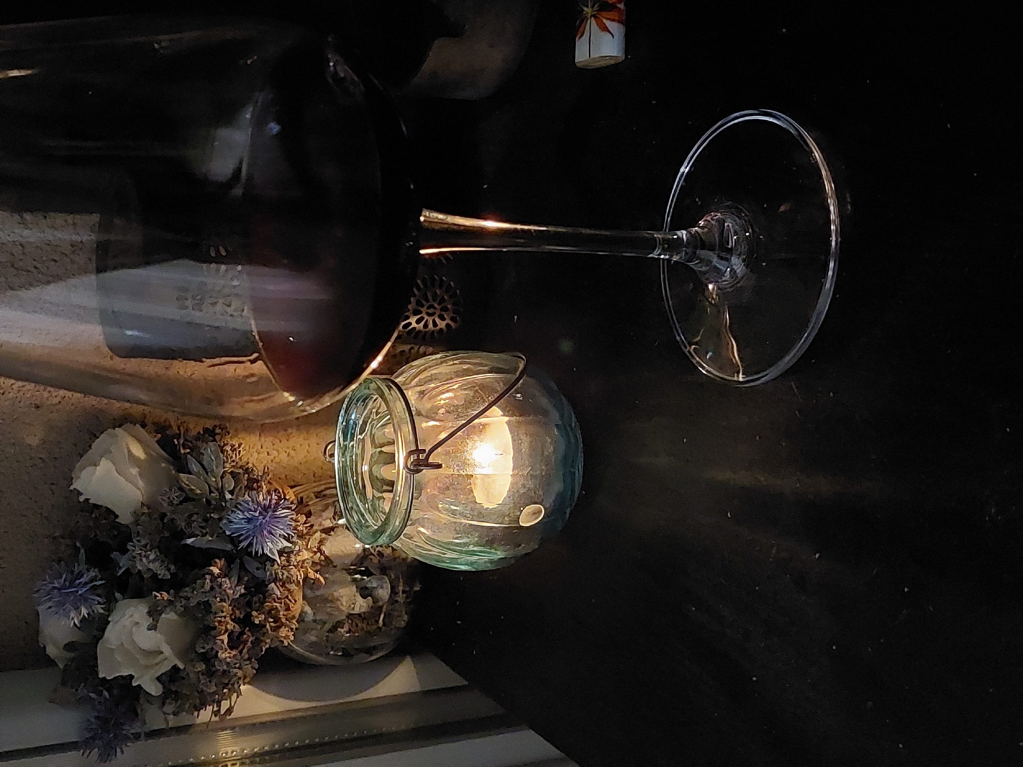 The image depicts an indoor still life scene under low-light conditions. No individuals are present.
**Location:** The specific location within the city of Plovdiv, Bulgaria, cannot be determined from the visual information provided in the image. The scene is generic and lacks identifiable landmarks or specific cultural cues.
**Subjects and Objects:**
The primary subjects are arranged on a dark, flat surface, possibly a table or shelf.
1. **Wine Glass:** A large, clear stemmed wine glass, partially filled with a dark red liquid, occupies the right foreground. Reflections from a nearby light source are visible on its stem and base.
2. **Glass Lantern with Candle:** To the left of the wine glass, a light teal-green, fluted glass lantern houses a lit, white tea light candle. The candle emits a warm glow, which is the dominant light source in the scene. A dark metal handle is attached to the top rim of the lantern.
3. **Flower Arrangement:** In the upper left background, a vase contains a dried or artificial floral arrangement. This arrangement consists of three off-white roses and several spiky, purple-blue thistle-like flowers, intermingled with desiccated brown and grey foliage. The vase appears to be reflective.
4. **Background Elements:** Behind the wine glass, a dark, irregularly shaped object (possibly a container or vase) is partially visible. A small, rolled-up item with an orange and white pattern (possibly paper or a matchbook) is discernable on the far right edge of the surface. A metallic, circular, gear-like or floral-patterned decorative element is situated behind the stem of the wine glass. The background wall has a textured, neutral-toned surface. The surface supporting the objects appears dark and exhibits small specks, consistent with dust or minor debris.
**Lighting and Environment:**
The scene is dimly lit, with the candle in the lantern serving as the main illuminant. This creates areas of strong contrast, highlighting the glass objects and the candle flame while leaving much of the background and the liquid in the wine glass in deep shadow. The overall environment is indoors, characterized by a muted color palette dominated by dark tones, soft whites, and subtle blues and greens, indicative of a cozy or intimate setting.