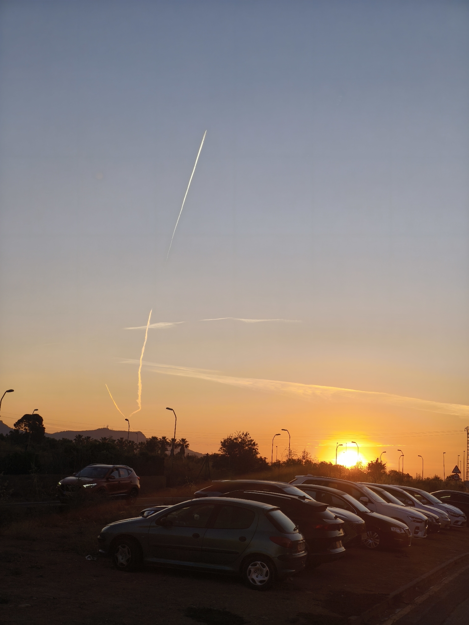 The image captures a parking lot at sunset in Nàquera / Náquera, Spain. Multiple cars are parked in rows, silhouetted against the setting sun. The sky transitions from blue to orange hues. Contrails from aircraft crisscross the sky. Distant trees and streetlights punctuate the horizon.
