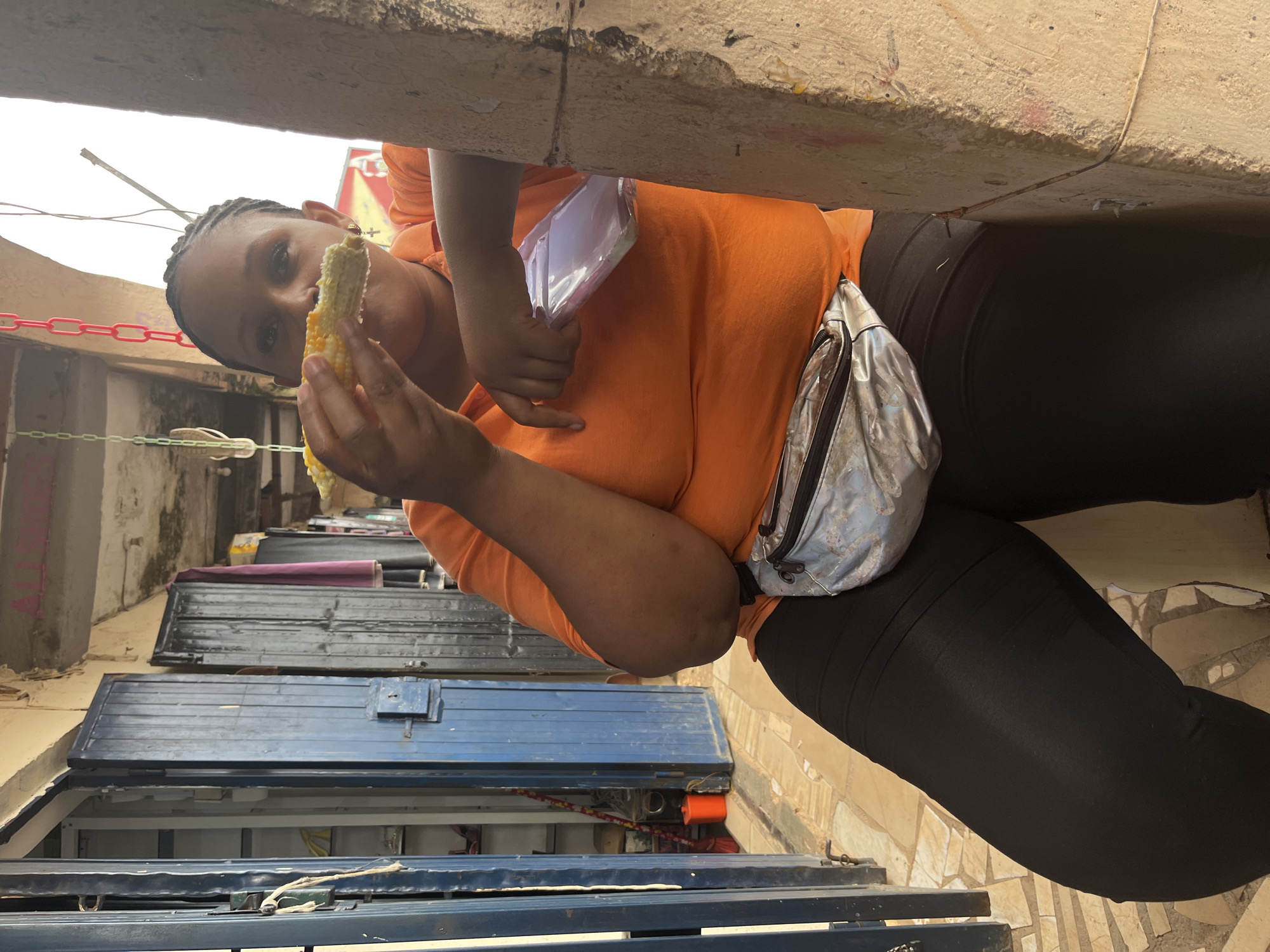 A person is holding and eating a cob of corn, positioned in a way that suggests they are leaning on a textured, concrete ledge above. They are wearing an orange t-shirt and black leggings, with a silver fanny pack worn across their waist. The background features stacked, blue and black metallic structures, possibly market stalls or storage units, with some wires and a red chain visible in the distance. The scene appears to be outdoors in a somewhat industrial or market setting. The person's expression is not clearly discernible due to the angle of the photo. The lighting suggests it could be daytime. The location context is Jos, Nigeria. There is no visible text in the image.