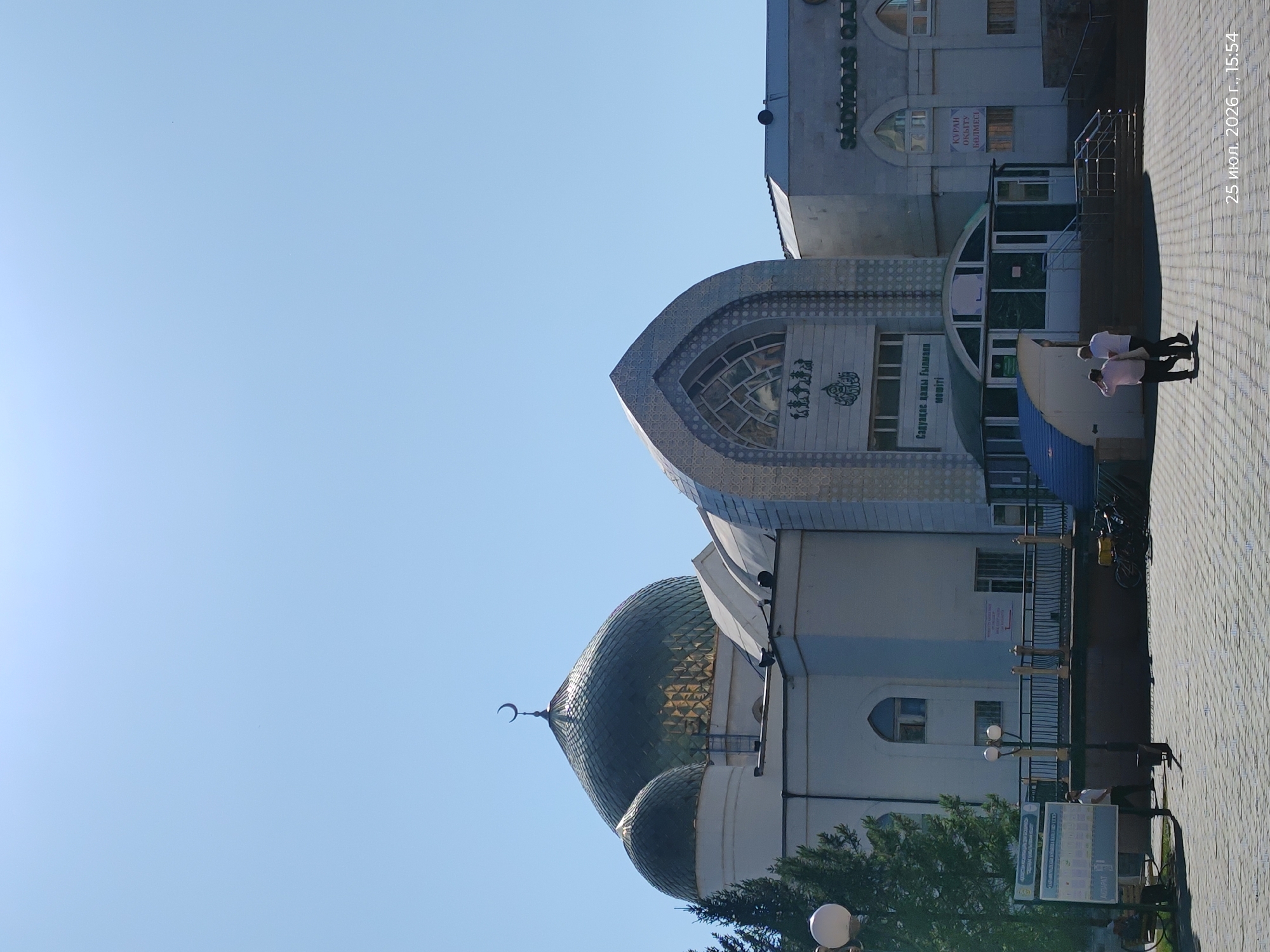 A street-level, full-shot photograph captures the exterior of a mosque in Astana, Kazakhstan, on a clear, sunny day. The mosque features a dominant large green dome topped with a crescent moon, and a smaller green dome to its left. The main building has a white facade, with intricate Islamic geometric patterns and Arabic script adorning the entrance. Adjacent to the main structure, a modern-looking building with arched windows displays signage in Cyrillic script, including "SADYNAS QIAY" and "КУРАН", indicating it may be related to Islamic education or community services. The foreground shows a paved walkway with a patterned stone surface. Two individuals, one in a pink shirt and dark pants, the other in a white shirt and dark pants, are walking away from the camera on the walkway. A bicycle with a yellow cargo box is parked to the right. In the background, a street lamp with two lights is visible. The date "25 июл. 2026 г., 15:54" is superimposed on the bottom right corner.
