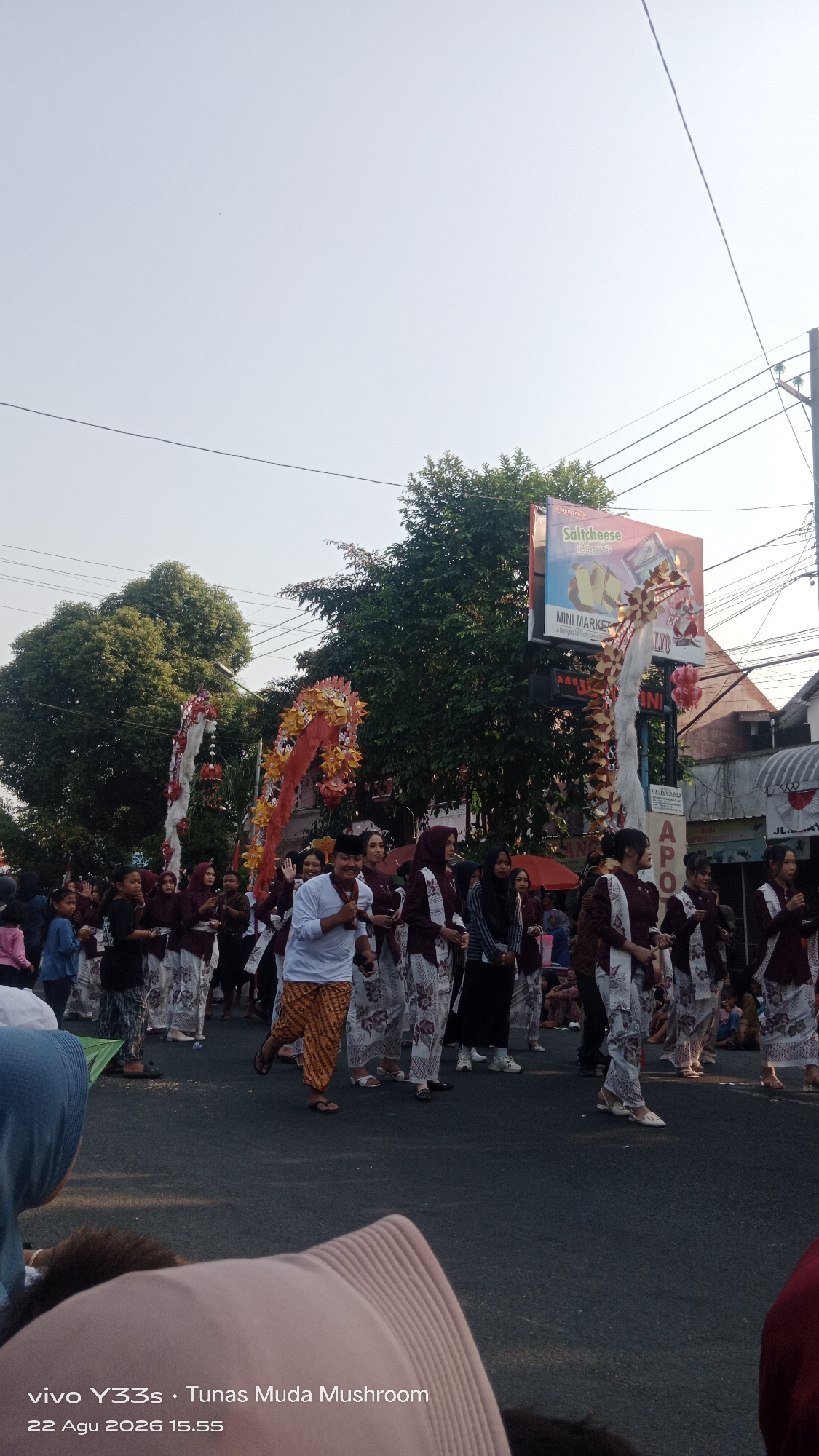 In Genengan, Indonesia, a group of people, primarily young women, are participating in a procession. They are dressed in traditional clothing, featuring dark maroon tops with ornate white and red patterned accents on the sleeves and hems, paired with white patterned sarongs. Some individuals are wearing headscarves.
In the foreground, a man in a white long-sleeved shirt and an orange and brown batik sarong is smiling and looking towards the camera, appearing to lead or be part of the parade. To his left, a group of girls in similar attire are also engaged in the procession.
The procession is taking place on a paved street. On the right side of the street, there are buildings, including one with a sign that reads "Saltcheese Crispy" and "MINI MARKET". Overhead, there are utility wires and poles. The background is dominated by trees and a clear, bright sky, suggesting it is daytime. The overall atmosphere appears festive, likely a cultural event or celebration. The timestamp "22 Agu 2026 15.55" and "vivo Y33s · Tunas Muda Mushroom" are overlaid on the bottom left of the image.