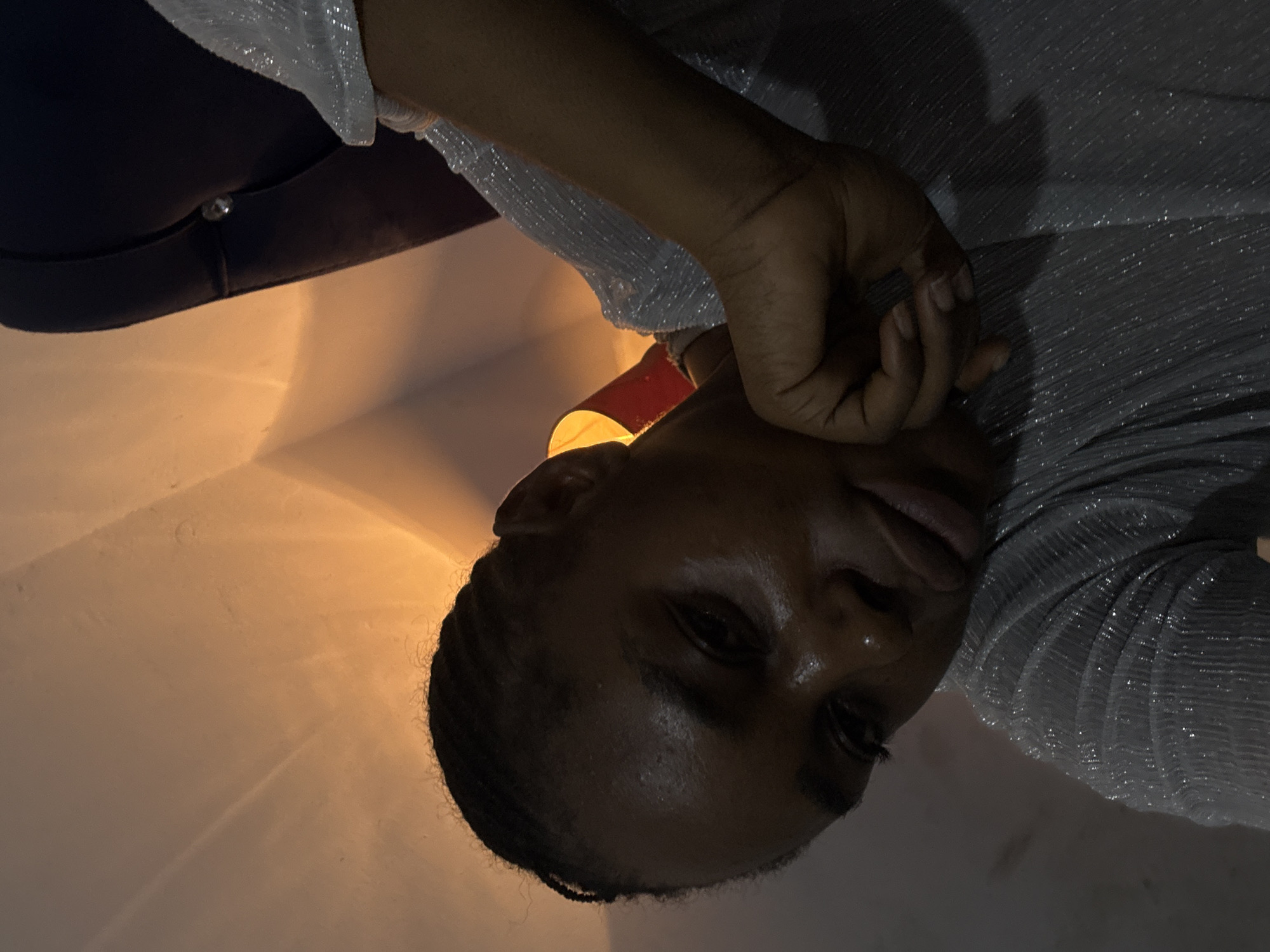 A person is depicted indoors, likely at night, as suggested by the dim lighting. The individual has dark skin and dark hair, possibly styled in braids or close to the head. They are wearing a shiny or textured shirt or garment. Their face is tilted towards the camera with their hand near their face. A dark colored chair is partially visible. The light source appears to be warm. The location cannot be determined from this image alone. It is impossible to confirm the image was taken in Abuja, Nigeria.