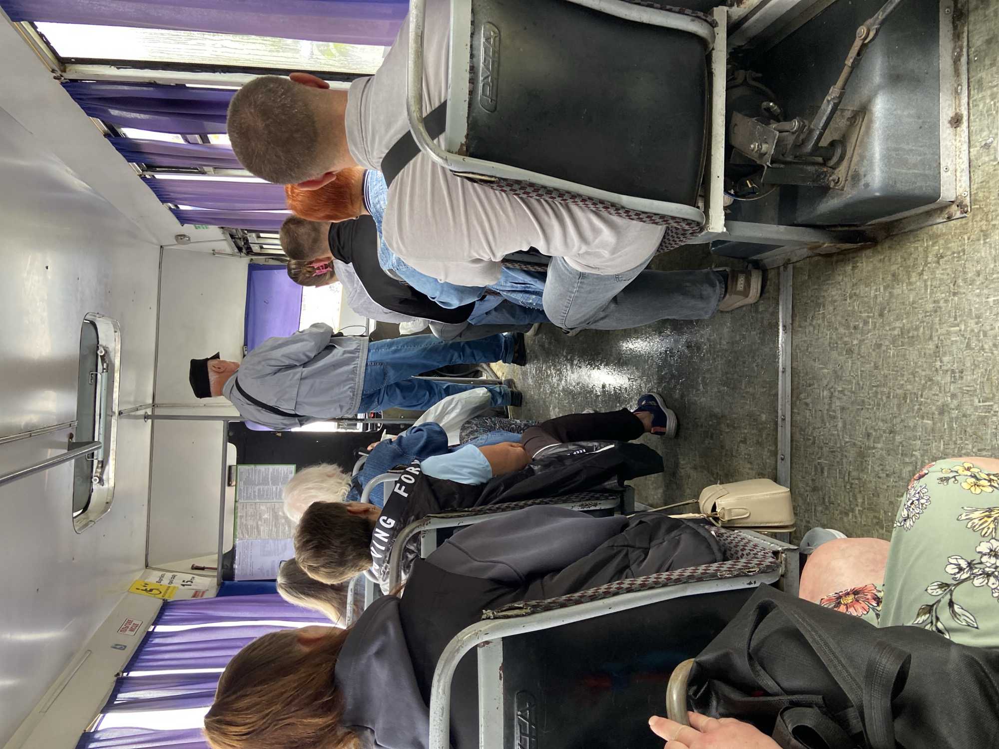 The image shows the interior of a bus in Nyzhni Mlyny, Ukraine. Several passengers are seated, facing forward. The bus has purple curtains along the windows. The floor appears wet. A man in a gray jacket and black hat stands in the aisle, facing away from the camera. The overall scene depicts a typical moment on public transportation.