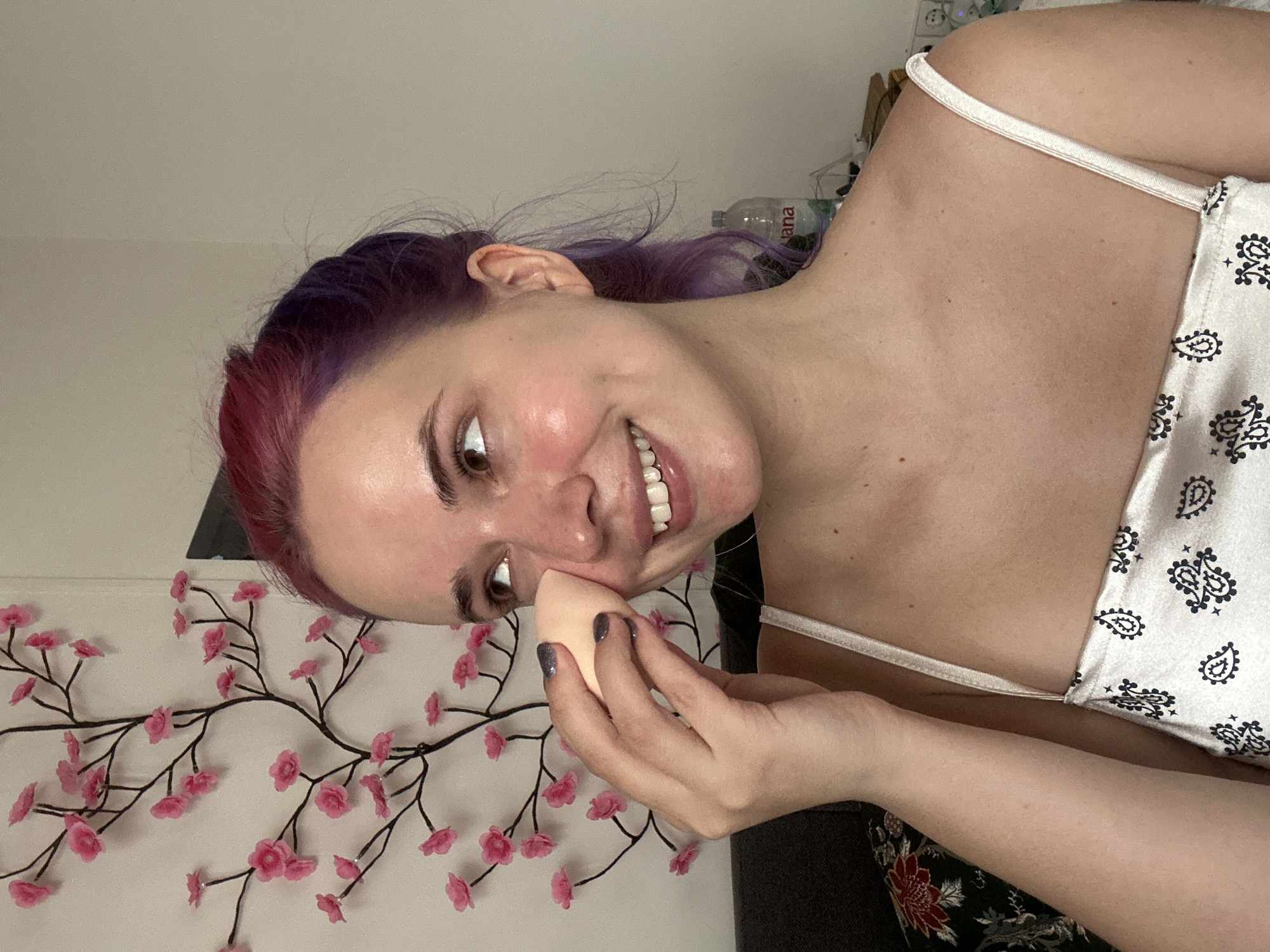 A woman is applying makeup with a sponge. Her hair is dyed purple and red. She is wearing a white top with a paisley pattern. There is a decorative floral branch in the background. A water bottle labeled "Jana" is visible behind her head. The location is the City of Zagreb, Croatia, based on the "Jana" water bottle, which is a Croatian brand.