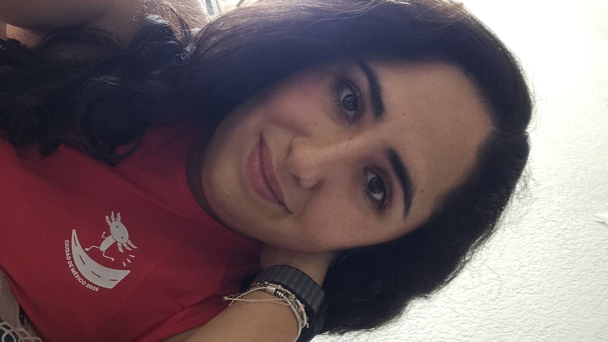 A young woman is shown in a selfie. She has dark hair, light skin with freckles, and is wearing a red t-shirt with a white logo that says "Ciudad de Mexico 2026". She is wearing a watch and bracelets on her left wrist. The background is a textured white wall. There are no indications of San Bernardino Tlaxcalancingo.
