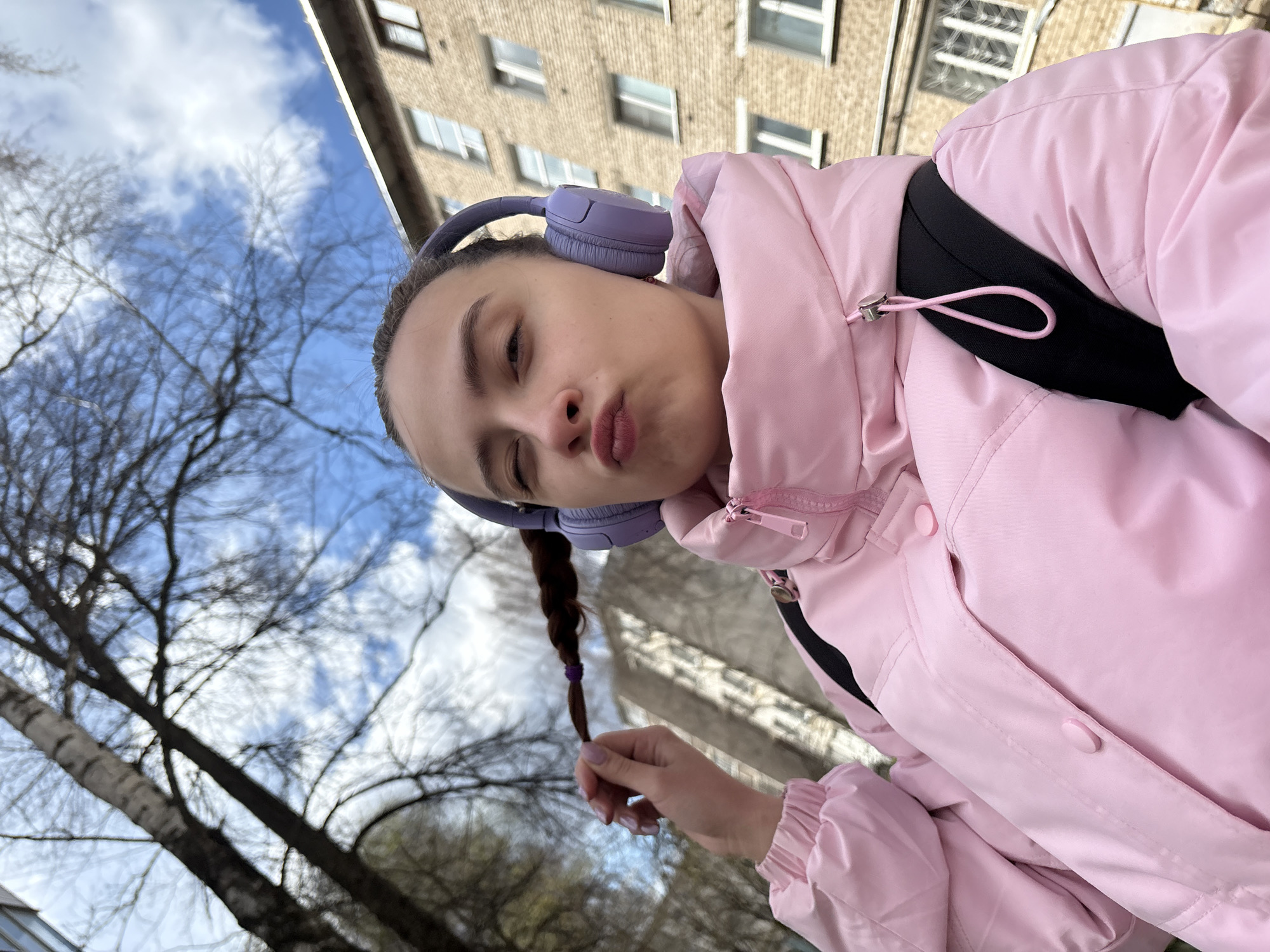 A young female is captured in a selfie outdoors, holding one of her braided pigtails with her right hand and making a playful expression with slightly puckered lips. She wears large, purple over-ear headphones and a light pink puffer jacket, with a black strap from what appears to be a backpack visible over her left shoulder.
The image is taken from a low angle, looking upwards. In the background, a multi-story apartment building constructed from light-colored bricks or stone with multiple rectangular windows is visible. Bare tree branches are prominent against a bright blue sky dotted with white clouds, indicating a late autumn, winter, or early spring setting with cool, clear weather. The overall scene suggests a daytime setting in an urban or suburban environment, consistent with the provided location context of Chaikovsky, Russia. No visible text is present in the image.