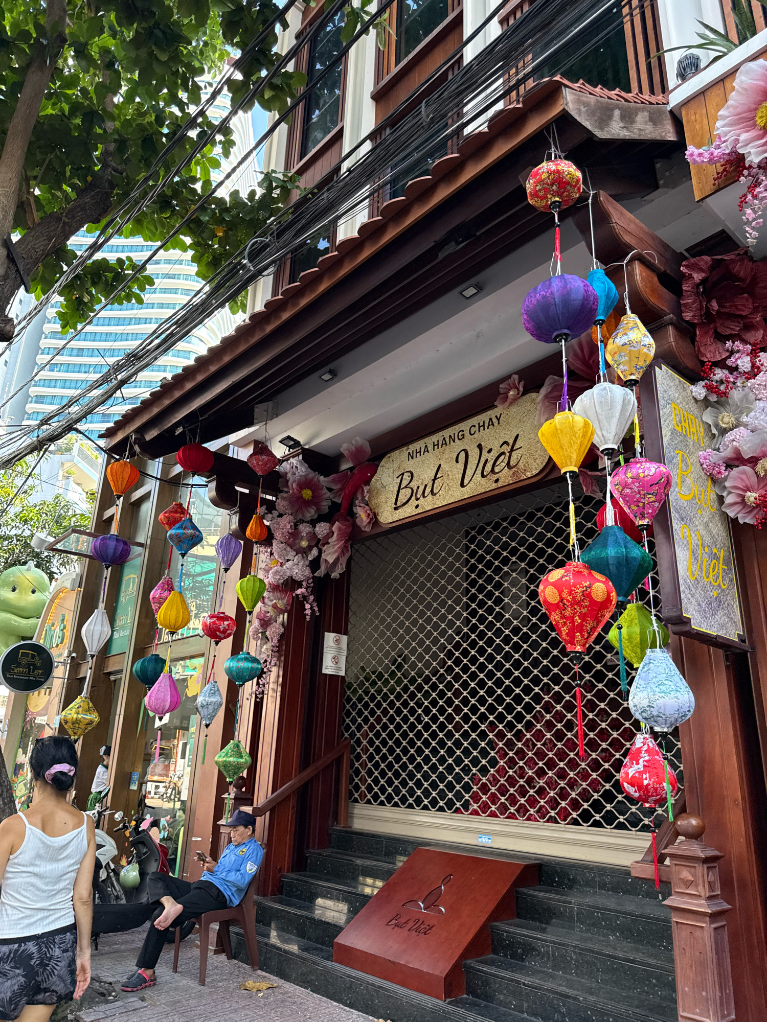 The image shows the exterior of a restaurant called "But Viet" in Nha Trang, Vietnam, on a sunny day. The restaurant features a traditional Vietnamese architectural style with a dark wooden awning adorned with numerous colorful, hanging lanterns of various shapes and sizes. The facade is made of dark wood, and the entrance is covered by a metal security grate.
In front of the restaurant, a man wearing a blue shirt and a dark cap is seated on a brown plastic chair, looking at his phone. He has bare feet. To the left of the man, a woman with dark hair tied back with a pink scrunchie is walking away from the camera. She is wearing a white tank top and dark patterned shorts. Various motorcycles are parked nearby.
Above the restaurant, a lush green tree with vibrant leaves is visible, with a tangle of electrical wires running across the frame. In the background, a tall, modern building with a distinctive curved facade and blue-tinted windows can be seen, indicating the urban setting of Nha Trang. The overall scene captures a slice of daily life in a Vietnamese city, blending traditional and modern elements.
