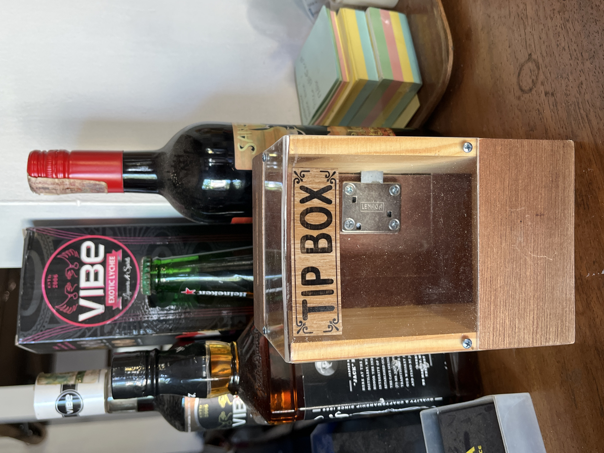 The image shows a close-up of a wooden tip box with a clear front panel and a metal lock. The words "TIP BOX" are carved into a wooden panel above the lock. Behind the tip box, several bottles of liquor are visible, including Jack Daniel's and a bottle of Vibe Lychee. A stack of colorful sticky notes is also present to the right of the tip box. The scene appears to be set on a wooden countertop or table. The image does not provide any information about the location in Karimunjawa, Indonesia, or the presence of any people or interactions.
