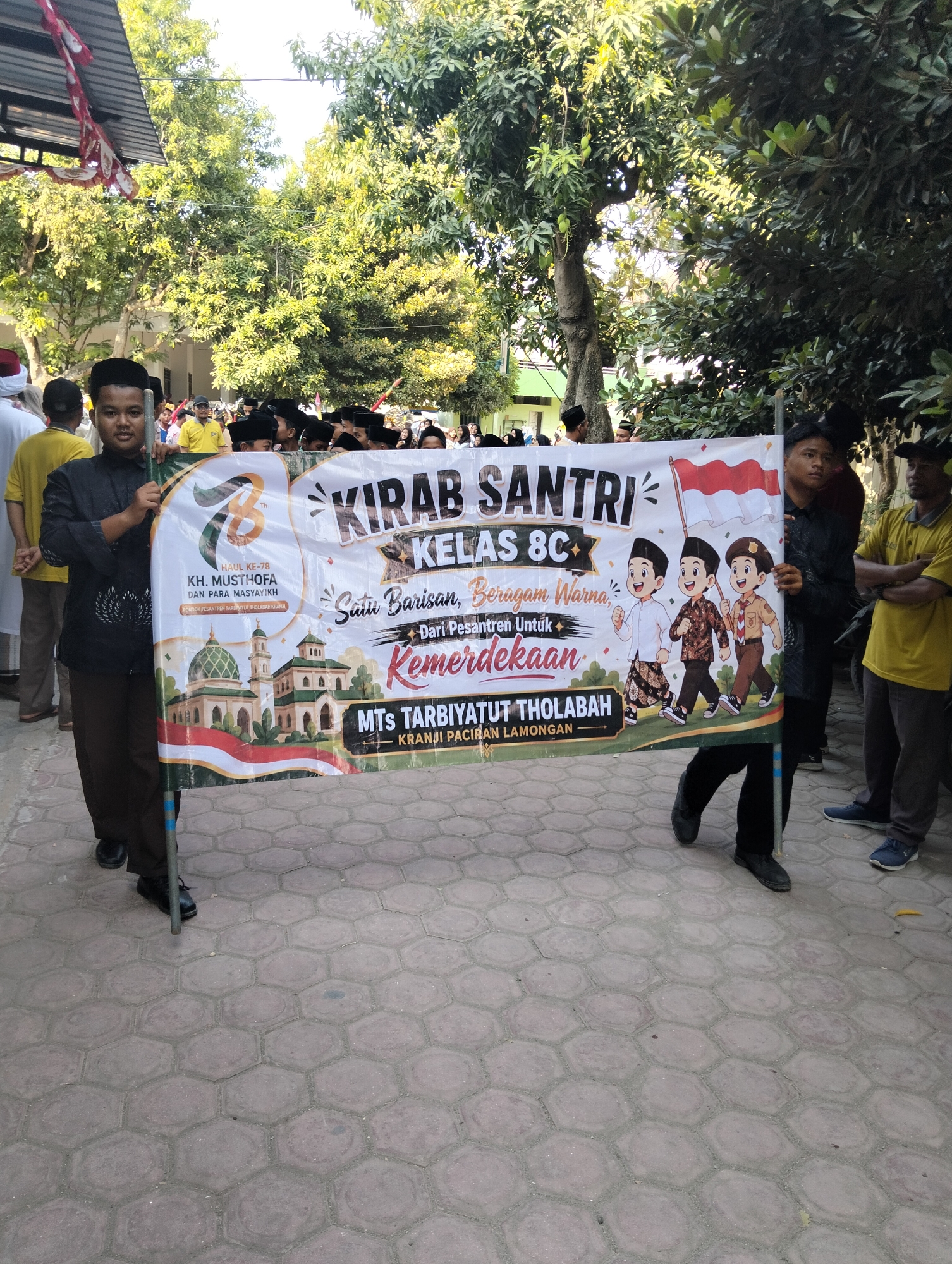 A group of young Indonesian boys are participating in a parade, holding a large banner that reads "KIRAB SANTRI KELAS 8C". The banner also features illustrations of three cartoon characters dressed in traditional Indonesian attire, a mosque, and the Indonesian flag. The boys are wearing black peci hats and dark clothing. The parade appears to be taking place in a paved outdoor area, possibly a schoolyard, with trees and buildings visible in the background. The scene suggests a cultural or religious event, likely in Kranji, Indonesia, given the text on the banner referencing "KRANJI PACIRAN LAMONGAN".