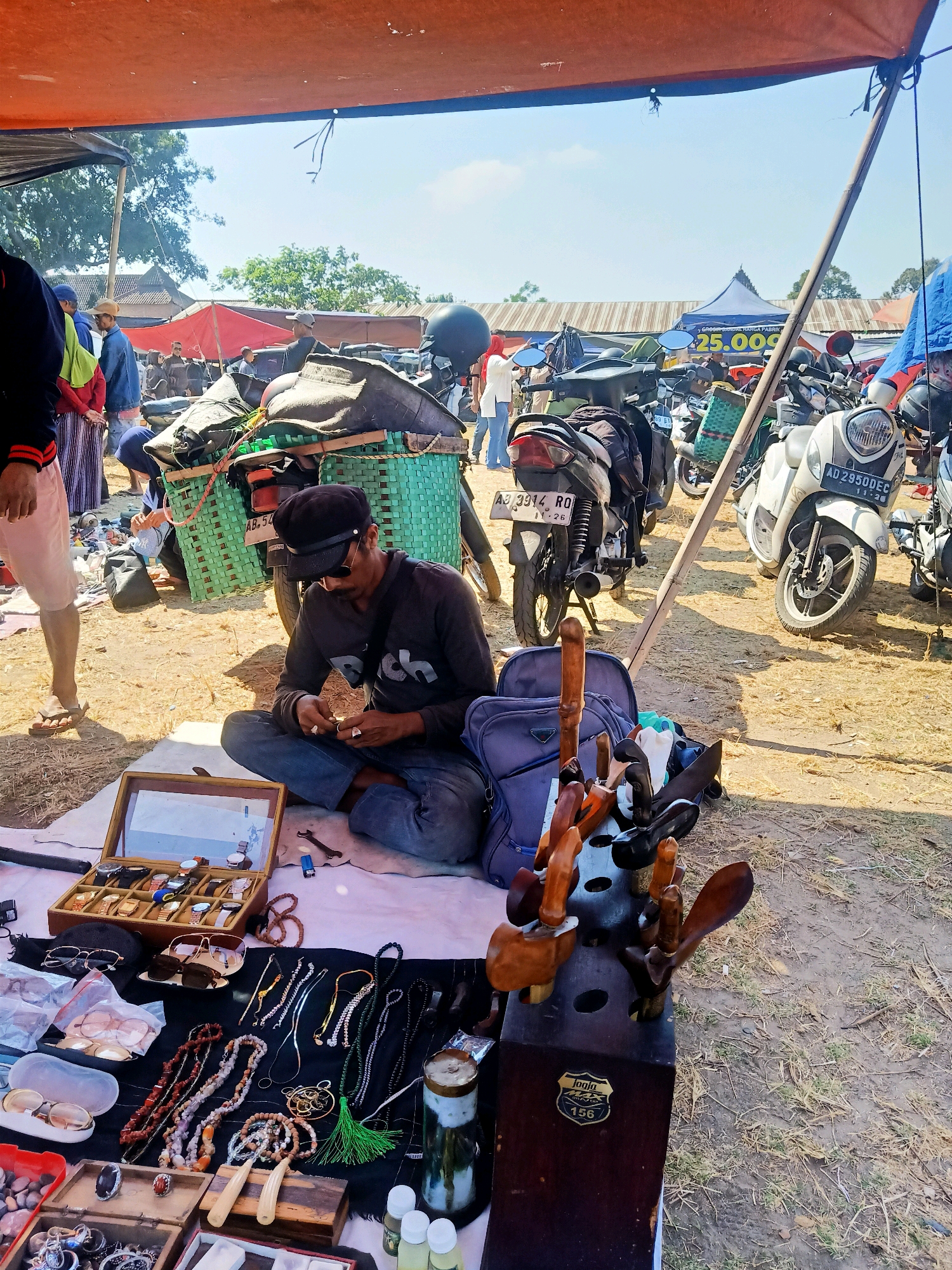 In Jatinom, Indonesia, a man sits cross-legged on a white cloth, tending to his wares at an outdoor market. The scene, under a large orange awning, is filled with various items for sale, laid out on the ground. The man, wearing a dark long-sleeved shirt, blue jeans, and a black cap, is focused on a small wooden box containing an assortment of watches. He holds a small object in his hands, appearing to be examining or repairing it.
Around him, the market buzzes with activity. A green woven basket overflowing with items is attached to the back of a motorcycle parked nearby. Other motorcycles are visible in the background, suggesting a busy commercial area. Tents and stalls create a colorful backdrop, with the sky a clear, bright blue.
The display of goods includes a variety of jewelry and accessories. A wooden box holds several sunglasses, while another section of the cloth showcases necklaces, bracelets, and rings. A wooden stand with carved wooden handles, possibly for tools or decorative items, is prominently placed in the foreground. Small bottles of liquid, possibly oils or perfumes, are also part of the merchandise. The ground is covered with dry grass and dirt, indicating an unpaved market area.