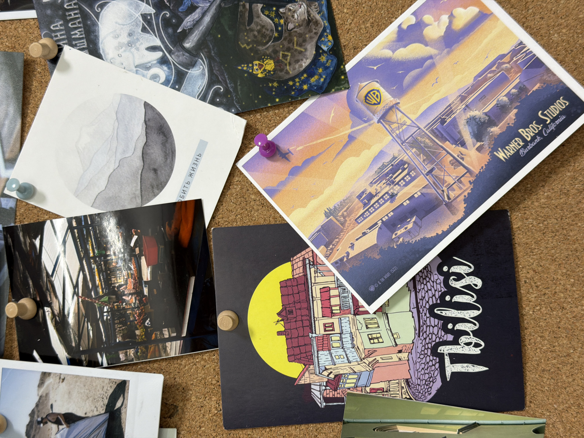 The image shows a corkboard with several pinned postcards and photos. One postcard features a stylized illustration of the Warner Bros. Studios in Burbank, California, highlighting the water tower with the WB logo. Another postcard depicts a colorful building against a yellow moon with the word "Tbilisi" written in script. Additional images include an interior shot of a market, a landscape with a round gray-toned illustration, and a dark illustration of a cat and owl. The corkboard is a neutral brown color, and the pins holding the images in place are visible.