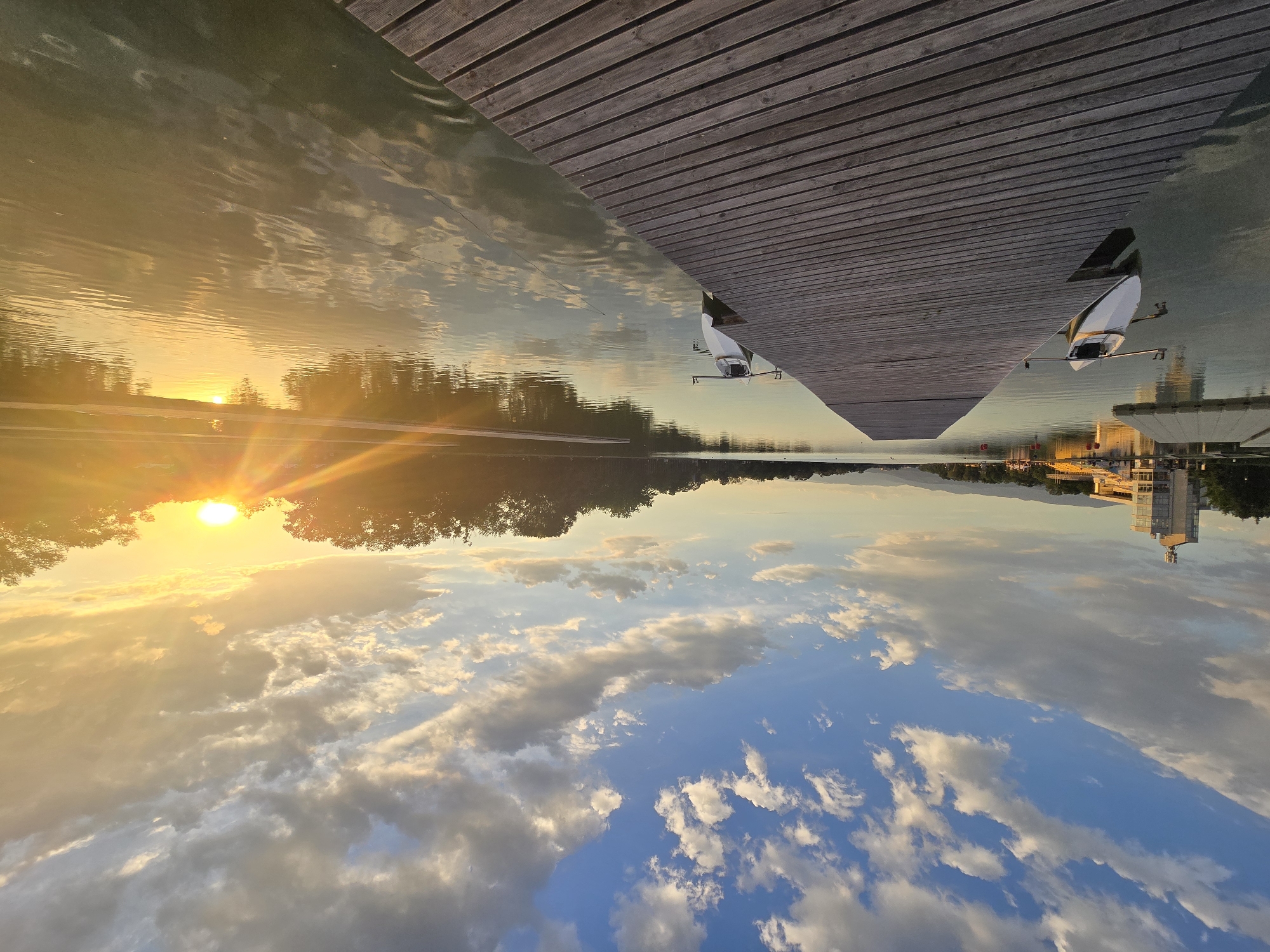The image shows a landscape with a wooden pier in the top right corner. In the water, there is a reflection of the sky with clouds. Across the water, there's a line of trees and a sunset. On the right, reflected in the water, is a tall building. There also seem to be a few boats docked on the right. The exact city is not identified.