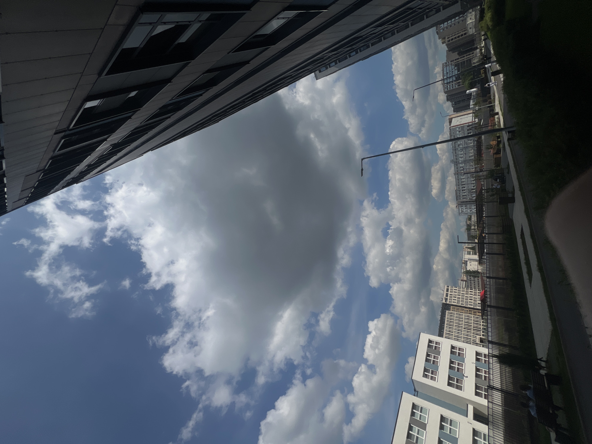A slightly angled, full shot captures a modern cityscape under a cloudy blue sky. The image is dominated by the rightward diagonal line of a tall, dark, modern building, its windows reflecting the sky. To the left, several lower-rise, white and light-colored buildings with numerous windows form a stark contrast. A black metal fence runs across the middle ground, separating a paved walkway and grassy areas from the buildings. Two figures are seated on a bench in the foreground to the left. Streetlights are visible, and in the distance, more buildings of varying heights and architectural styles populate the horizon. The environment appears to be a newly developed urban area.