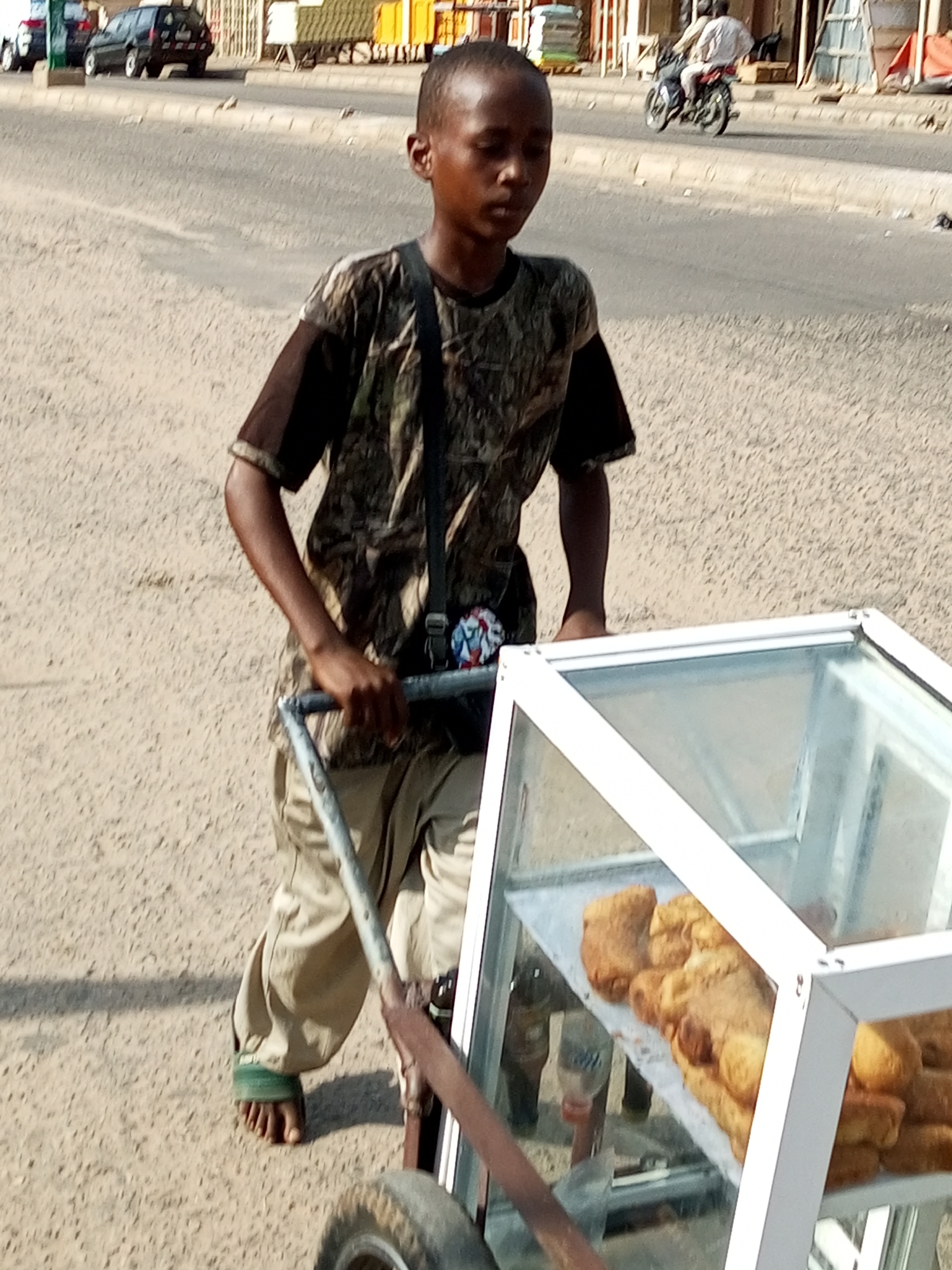 A young boy, appearing to be of East African descent, walks with a food cart on a dirt road in Arawa, Nigeria. The boy wears a camouflage t-shirt and tan pants, with a green band around his left ankle. He is pushing the food cart, which has a glass display case filled with what appear to be fried pastries. The cart has two wheels and a metal frame. In the background, cars and a motorcycle are visible on a paved road. The environment is an outdoor urban setting with a clear sky.