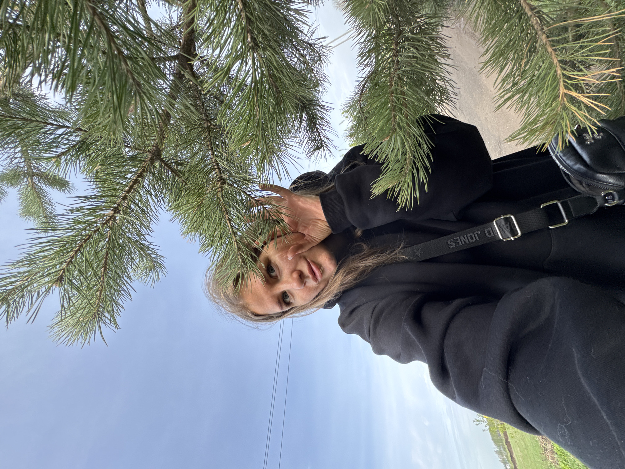 A young woman in a black hooded sweatshirt poses beneath pine branches against a sky in Горный, Russia. She holds her hand up near her face. A black shoulder bag is visible. In the background, fields and power lines can be seen.