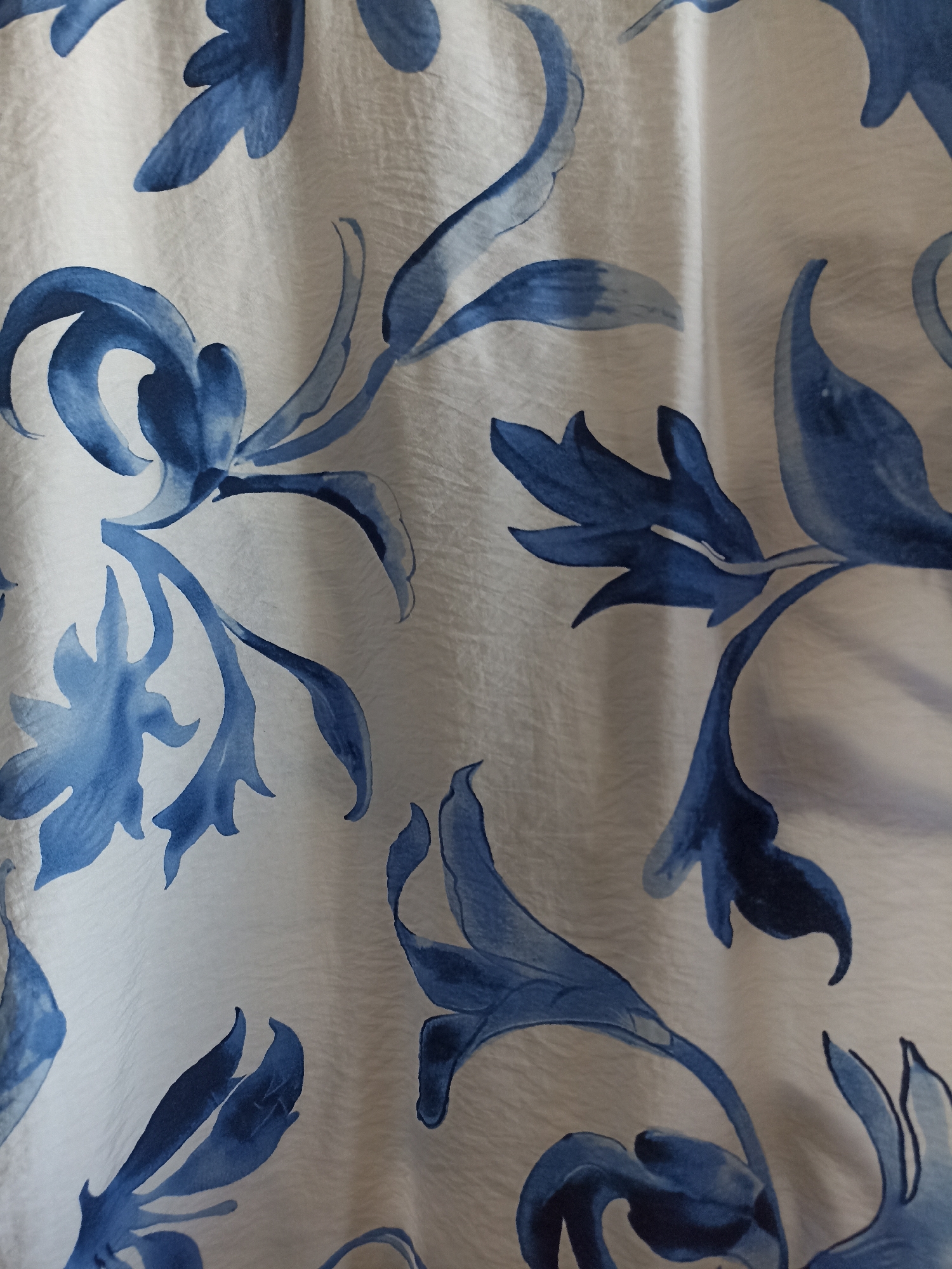 The image is a close-up, top-down view of a white fabric with a blue floral pattern. The pattern appears to be painted or printed in watercolor style, with flowing lines and varying shades of blue. The fabric has a textured surface, with visible wrinkles and folds that create shadows and highlights, giving it a slightly three-dimensional appearance. There are no people, animals, or other discernible objects in the image, only the fabric with its decorative pattern. The location in Novosibirsk, Russia, is not evident from the visual information provided.