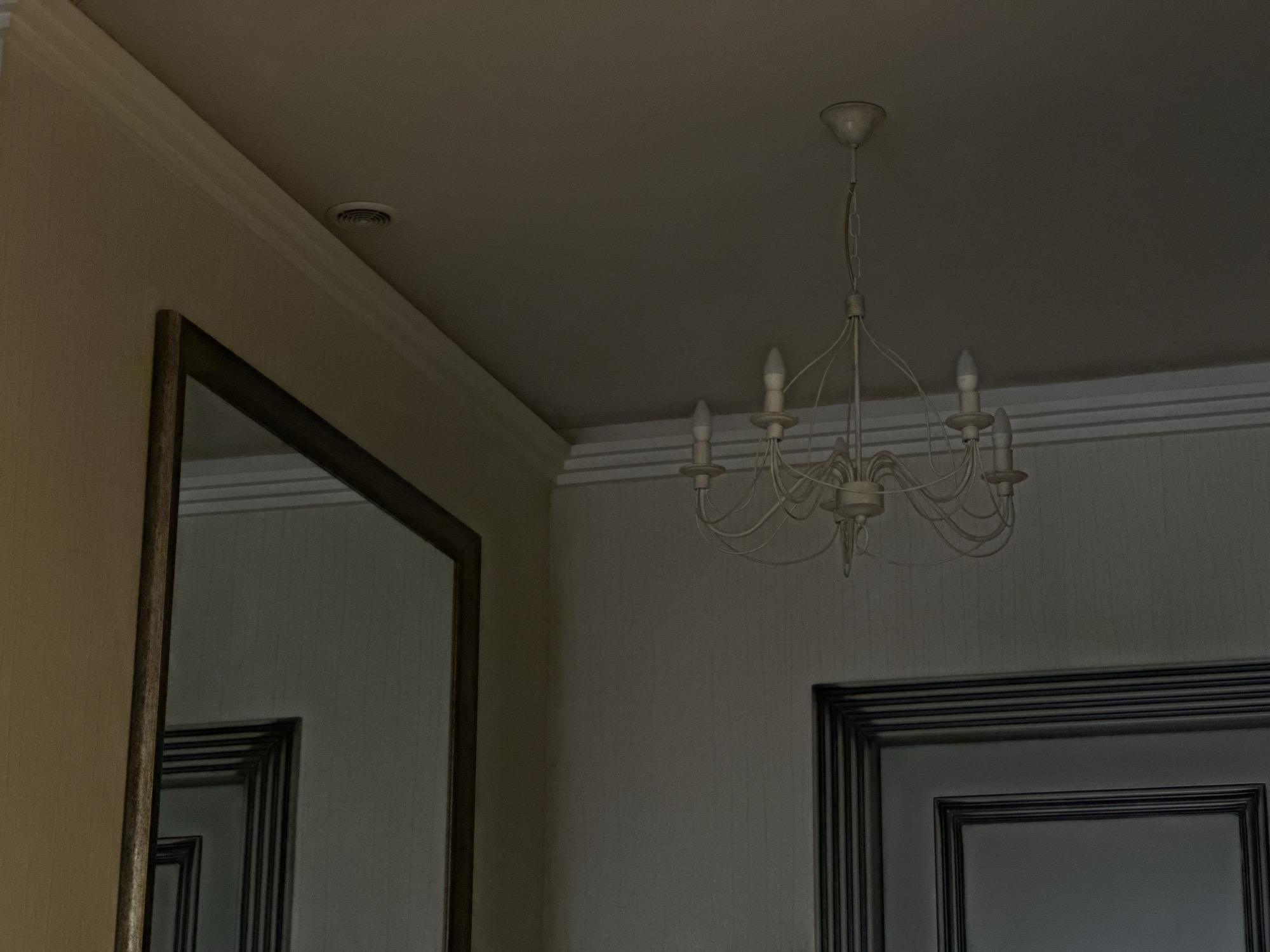 A dimly lit indoor shot captures a room interior, likely in Yerevan, Armenia, given the style of architecture and interior design. The scene features a mirror on the left wall, reflecting another framed element within it, giving an illusion of depth. On the right, a framed panel or door is visible, with a white chandelier dominating the upper-right part of the frame, suspended from the ceiling. A circular vent is near the upper-left corner.