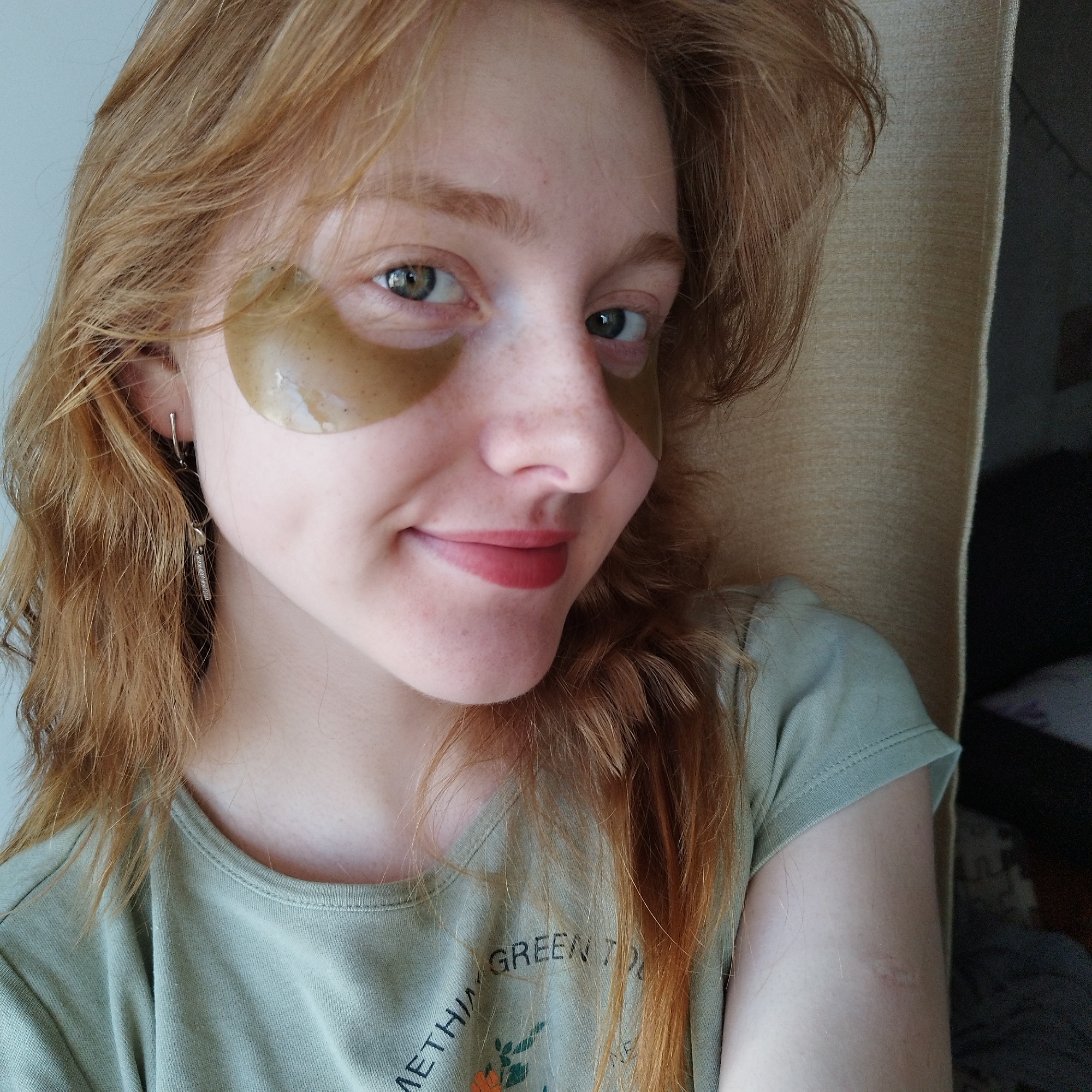 A young woman with long, wavy, reddish-blonde hair is captured in a close-up, eye-level portrait. She has fair skin, a light scattering of freckles across her nose and cheeks, and bright green eyes. She is smiling gently, with her lips painted a soft reddish-pink hue.
Underneath both eyes, she wears translucent, gold or amber-colored gel under-eye patches, which appear moist and reflective. Her hair falls around her face and shoulders. In her visible left ear, she wears two small, dangling silver earrings: one is a simple vertical bar, and the other features a small circular stud connected to a vertical bar with tiny textured details or rhinestones.
She is dressed in a light olive green short-sleeved t-shirt. Visible text on the front of her shirt reads "METHING GREEN T", with further text and a small graphic of an orange flower with green leaves beneath it. The phrase likely includes "SOMETHING GREEN".
The background is softly out of focus, showing a light blue-grey wall to her left and a textured, light beige fabric, possibly a curtain or furniture, to her right, with a darker, indistinct area beyond it. The lighting is bright and natural, suggesting an indoor, daytime setting. The woman appears relaxed and content, possibly engaged in a self-care routine.