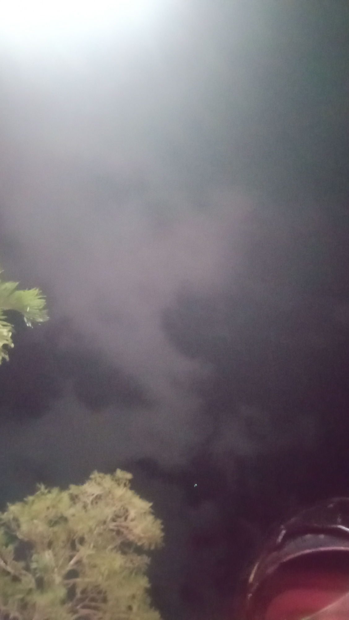 The image shows a night scene with a bright light source at the top left, casting a diffuse glow. Wispy clouds or smoke are visible in the upper and middle portions of the frame. In the lower left, the branches of a leafy tree are visible, appearing to be illuminated by the overhead light. In the lower right, a dark, possibly metallic object with a red accent is partially visible, suggesting a vehicle or helmet. The overall environment is dark, with the light source and the illuminated tree providing the main points of interest against a shadowy background. The location is identified as Kuta, Indonesia.