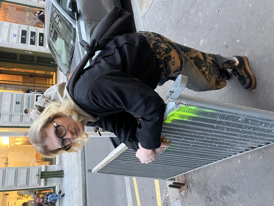 A person wearing glasses, a black jacket, and camouflage-patterned pants leans over a metal grate with a vibrant green stripe. The grate is on a city street with yellow lane markings. Behind the person, a silver car is parked next to a building with visible signs. Other pedestrians are walking in the background, adding to the urban scene. The person appears to be engaged with something on the grate, possibly interacting with it or retrieving something.