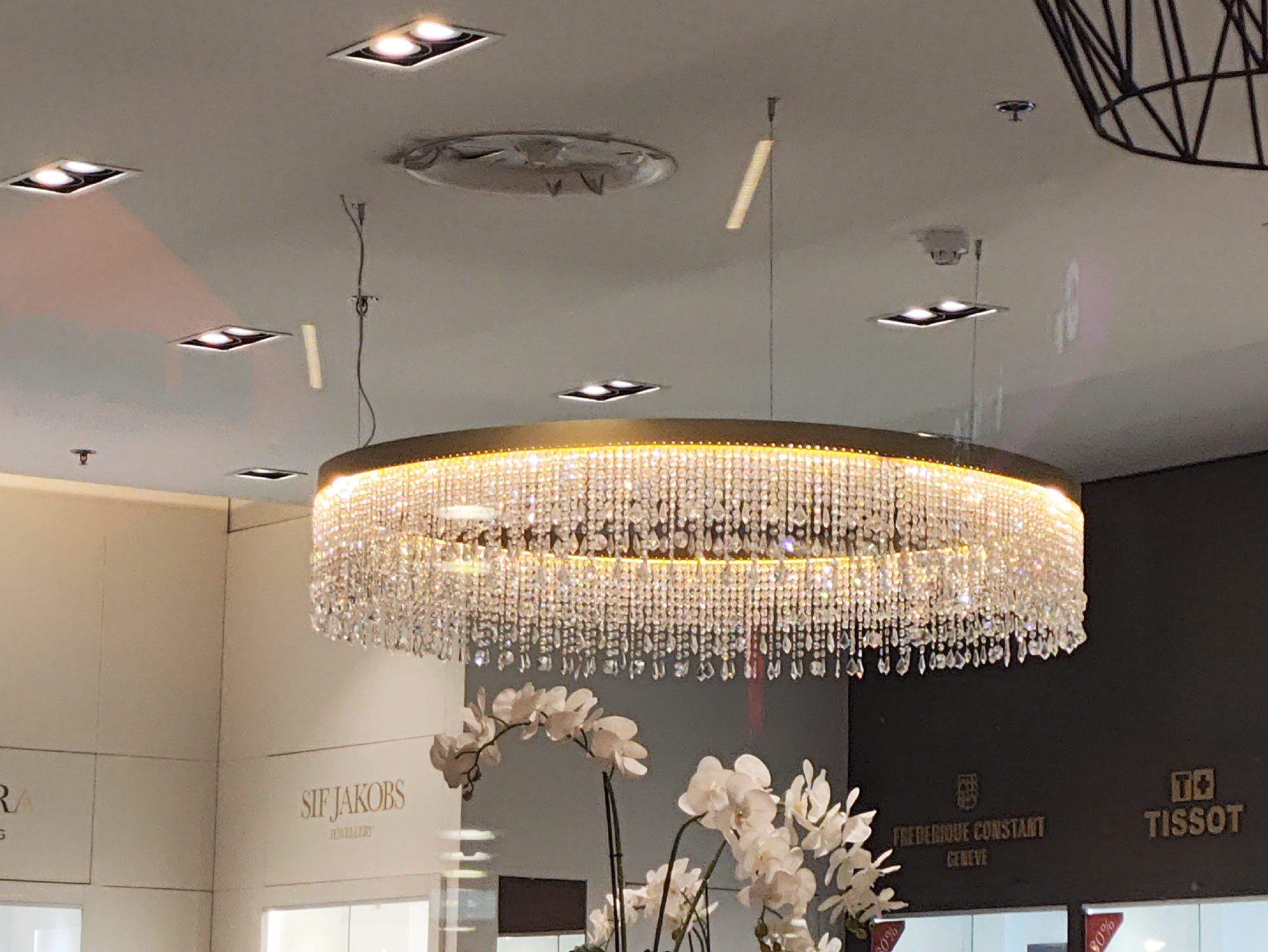 The image shows a large, circular crystal chandelier hanging from a white ceiling. The chandelier is adorned with numerous sparkling crystals and illuminated by an internal light source. In the background, there are several recessed square ceiling lights. To the left, a white wall displays signage for "SIF JAKOBS JEWELLERY." To the right, a dark wall features gold lettering for watch brands, specifically "FREDERIQUE CONSTANT GENEVA" and "TISSOT." White orchids are arranged in a planter in the foreground, partially obscuring the lower portion of the chandelier and the signage. The scene appears to be an interior shot of a jewelry or watch store. The location is identified as Tallinn, Estonia.