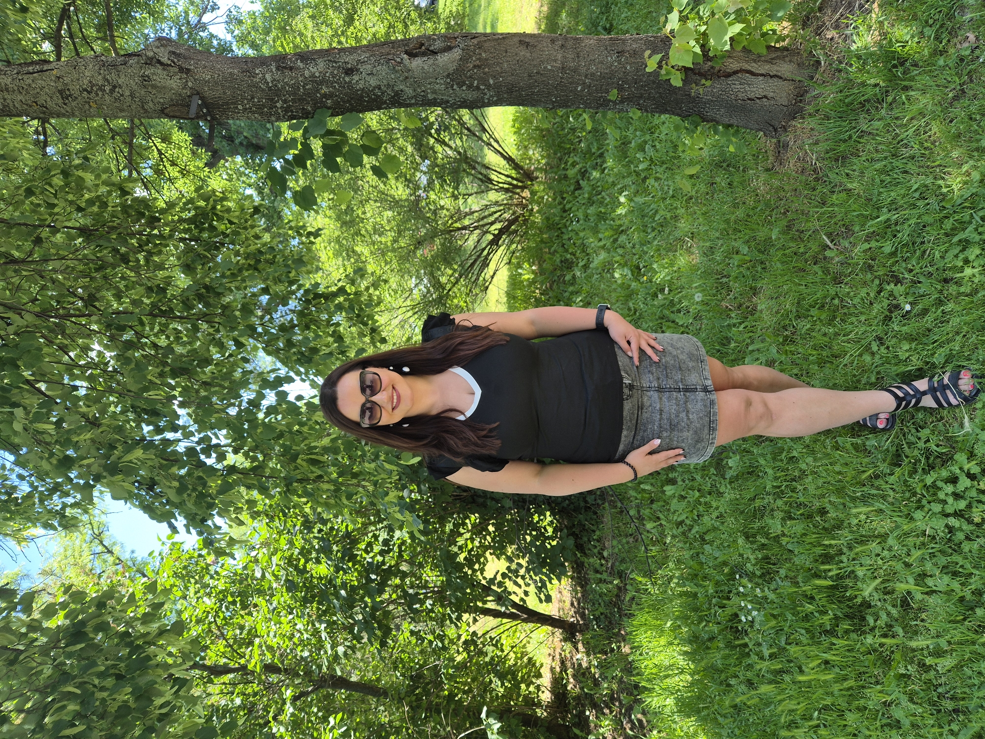 A medium-shot image shows a woman in an outdoor setting. She is lying on a grassy surface, positioned parallel to a tree trunk that is horizontally across the top third of the image. The woman is wearing sunglasses, a black t-shirt with a white neckline, and a grey denim skirt, with black sandals on her feet. Her dark hair is long and styled around her shoulders. The ground is covered in green grass and clover. The background is filled with lush green foliage and trees. There are no indicators to determine the specific location or city.