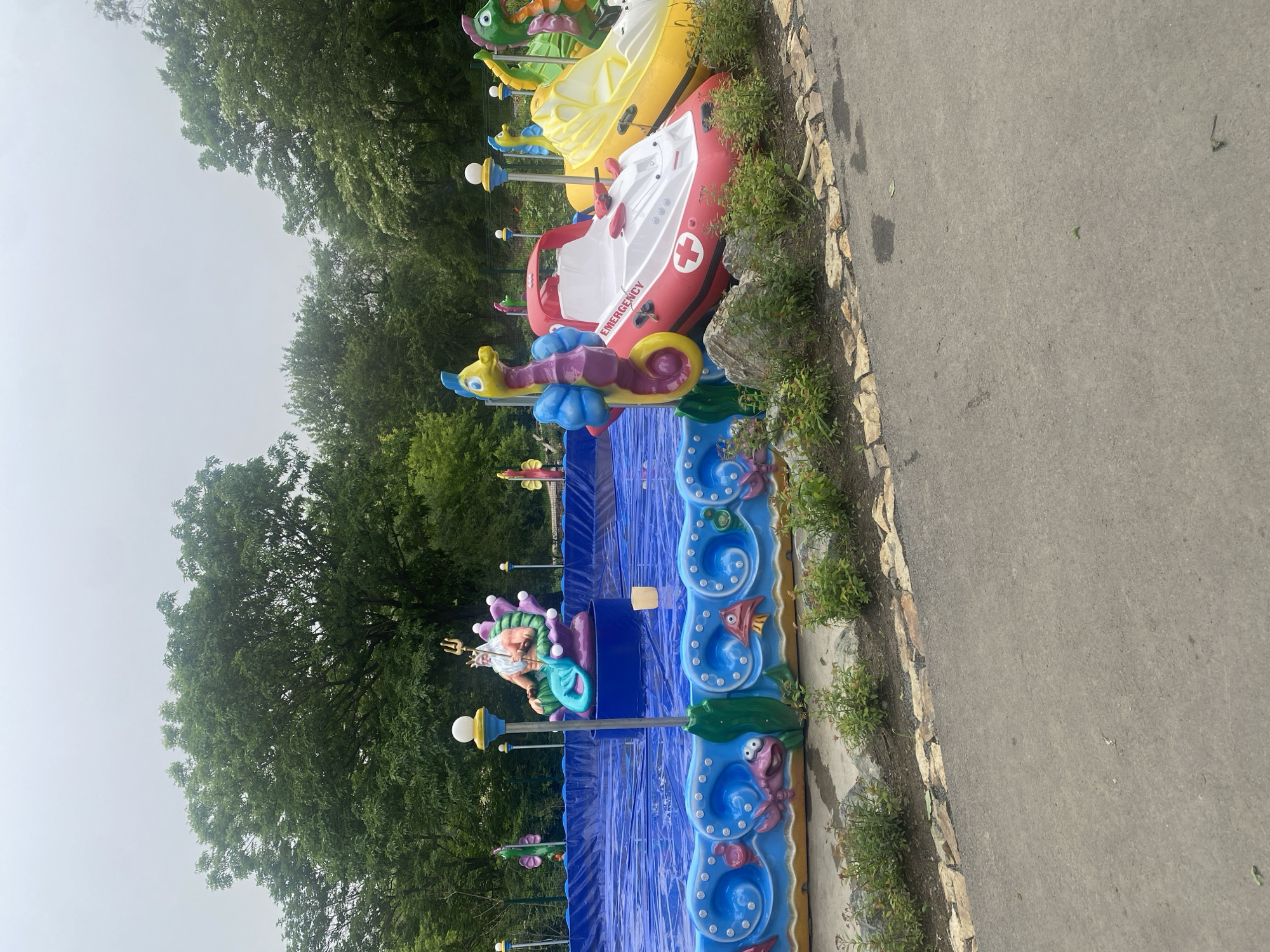 An outdoor amusement park ride features a pool with blue lining and decorative sea creatures along the edge. In the center of the pool is a statue of Neptune. Several brightly colored amusement ride vehicles are parked in a row along the edge of the pool, including a red and white boat with an emergency symbol, a yellow boat, and other sea-themed vehicles. The ride is surrounded by trees and a gray sky.