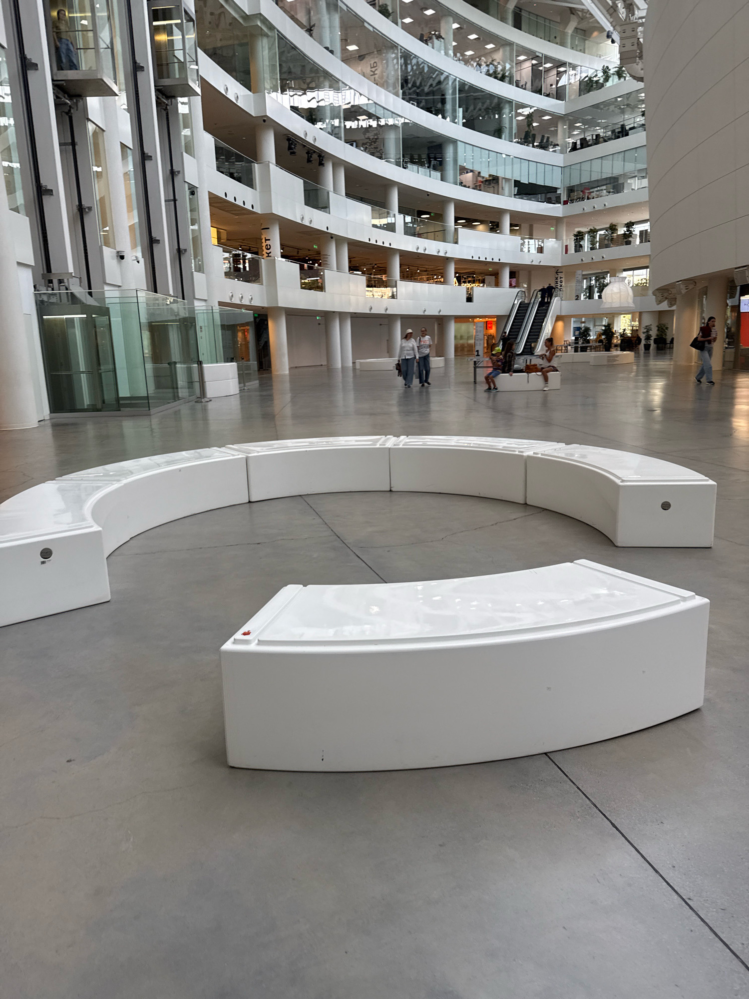 A wide shot captures the interior of a modern, multi-story atrium. White, modular benches, some arranged in a curve and others as individual units, are prominently featured in the foreground. The building's architecture is characterized by white facades, glass balconies overlooking the atrium, and exposed structural elements. Elevators in glass enclosures are visible on the left, and escalators lead to upper levels on the right. Several people are scattered throughout the space, some walking and others seated on benches. The floor is a polished concrete or stone surface. The overall impression is of a bustling public space within a contemporary architectural complex. Based on the signage visible, this is likely the Yeltsin Center in Yekaterinburg, Russia.