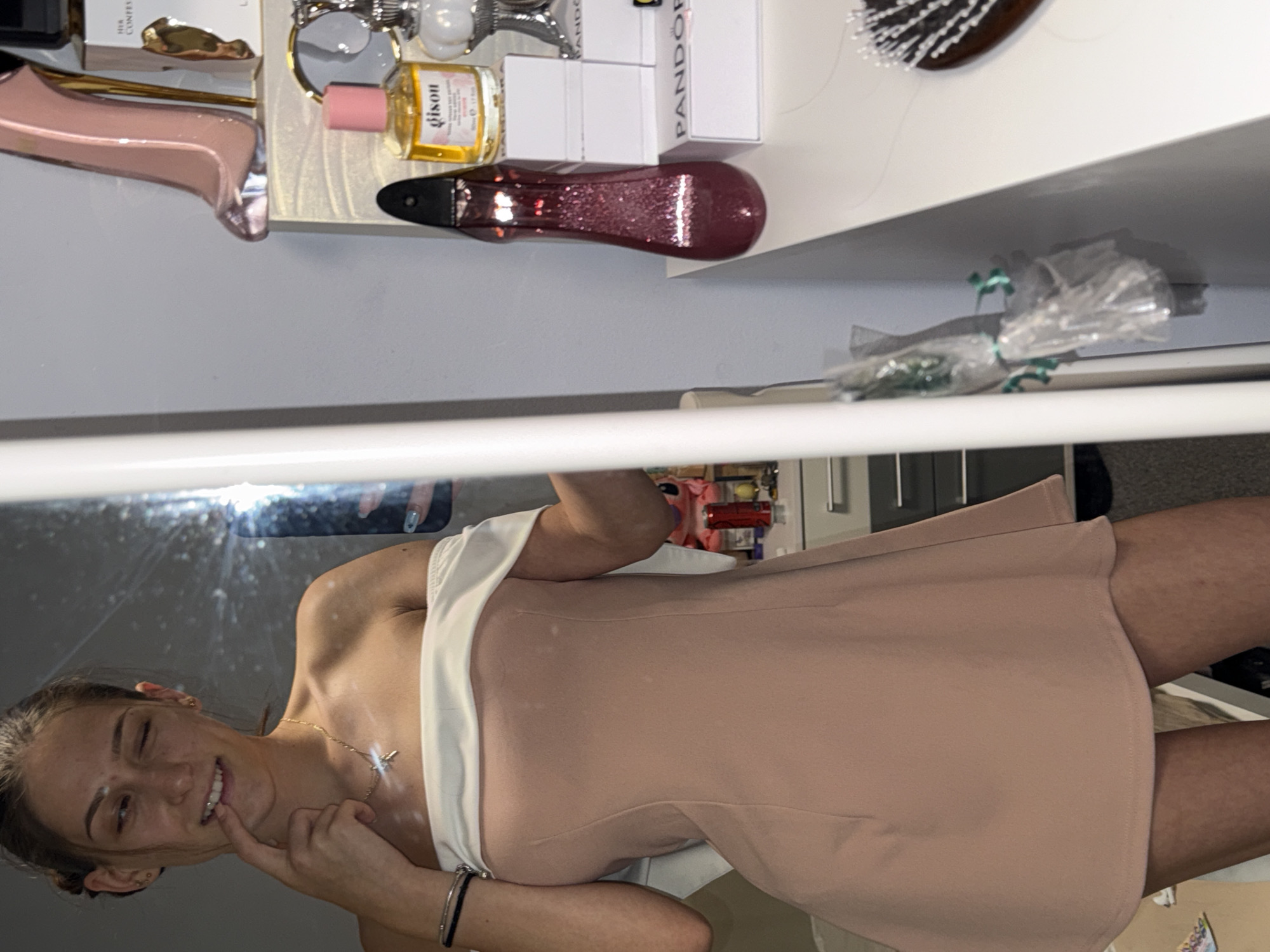 In an indoor setting, a young woman stands in front of a mirror, taking a selfie. She is wearing an off-the-shoulder, peach-colored dress and a delicate necklace. Her right index finger is touching her lips. The background reveals a dresser with various items, including perfume bottles, jewelry boxes, and a hairbrush. The lighting suggests a personal space, possibly a bedroom or dressing area, within a residence in Sofia, Bulgaria.