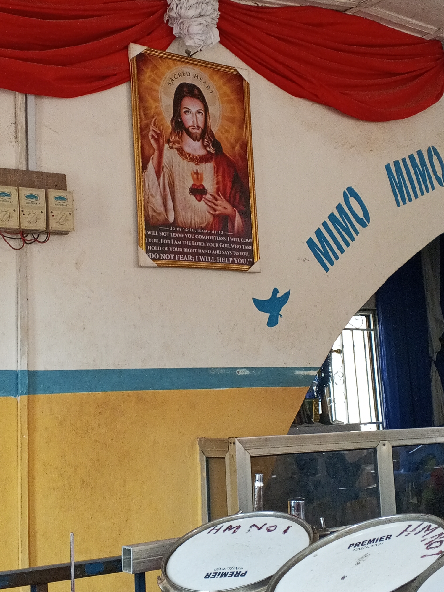 The image shows an interior scene in Lokoja, Nigeria. Dominating the composition is a wall with a blue and yellow horizontal band. Above this, a religious print depicting Jesus with the Sacred Heart hangs, draped with red fabric and a white flower-like ornament. To the right of the print, "MIMO MIMO" is stenciled in blue above an archway. A blue bird-shaped stencil is present below the text. Beneath the wall, drums with "PREMIER" brand and the hand-written text "HM No 1" are visible. The scene suggests a space associated with a church or religious organization. Electrical boxes labeled "ORIGINAL" are located on the left side of the wall. A doorway is visible through the archway. The overall impression is of a modest and aged interior.