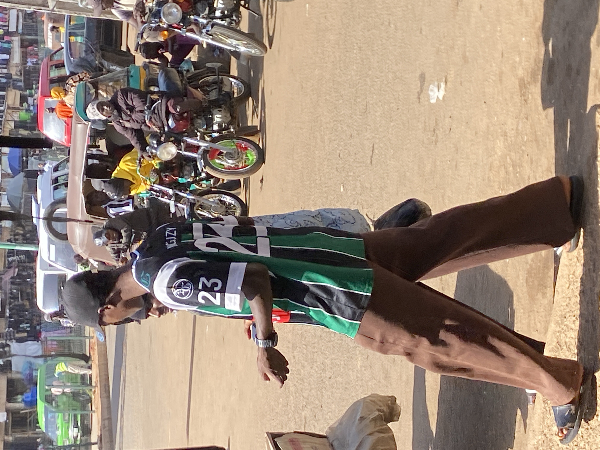 A man wearing a black baseball cap, a black and green sports jersey with the number 23 and the name "NGIZZY" on the back, and brown pants stands on a paved street. He wears a silver wristwatch on his left wrist and sandals on his feet. He is looking to his left with his left arm slightly raised.
Behind him is a street scene that includes a mix of pedestrians, parked motorcycles, and cars. Various buildings and street vendors line the background. Based on the presence of motorcycles and the overall crowded street environment, the photo was taken in Bukuru, Nigeria.