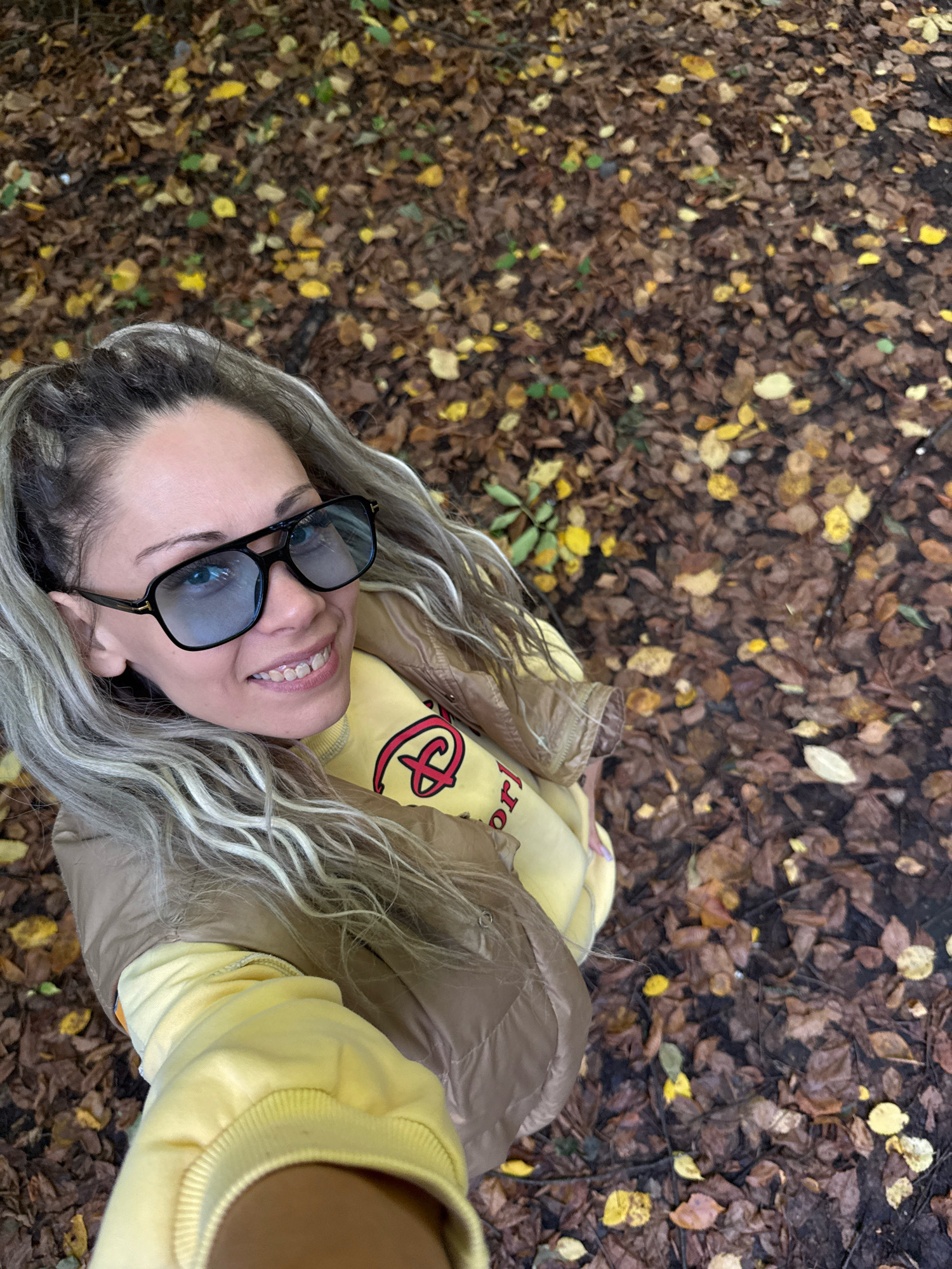 A woman is posing for a selfie outdoors, looking up at the camera with a smile. She has long, blonde hair pulled into a ponytail and is wearing large, dark-rimmed sunglasses. She is also wearing a yellow sweatshirt with a red design on it, and a brown puffer jacket. The background is a ground covered with fallen autumn leaves, suggesting a park or wooded area. The location is identified as Rusanovo, Russia.