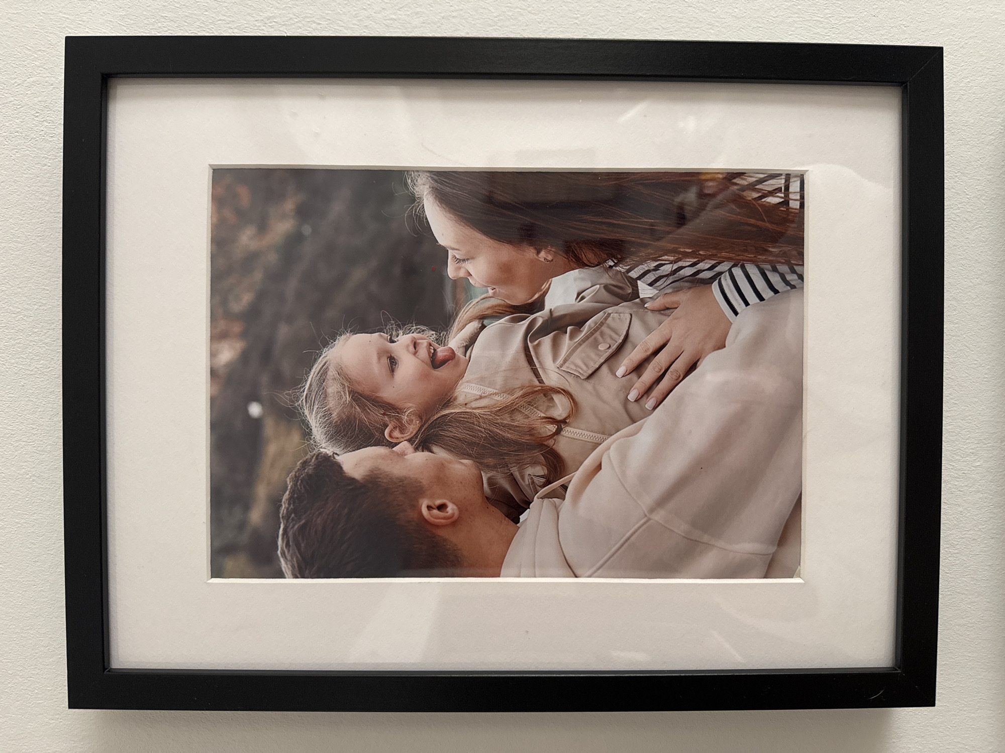 A framed photograph hangs on a white wall. The photograph depicts a family of three, consisting of a mother, father, and young daughter. The mother is on top, wearing a striped shirt and beige jacket, with her hand resting on the daughter's stomach. The daughter is lying on her back, sticking her tongue out playfully. The father is underneath, looking up towards his family. The background appears to be outdoors, possibly a park with trees or foliage. The photograph is mounted with a white mat board inside a black frame. The location of the photograph is unknown; it's just hanging on a wall.
