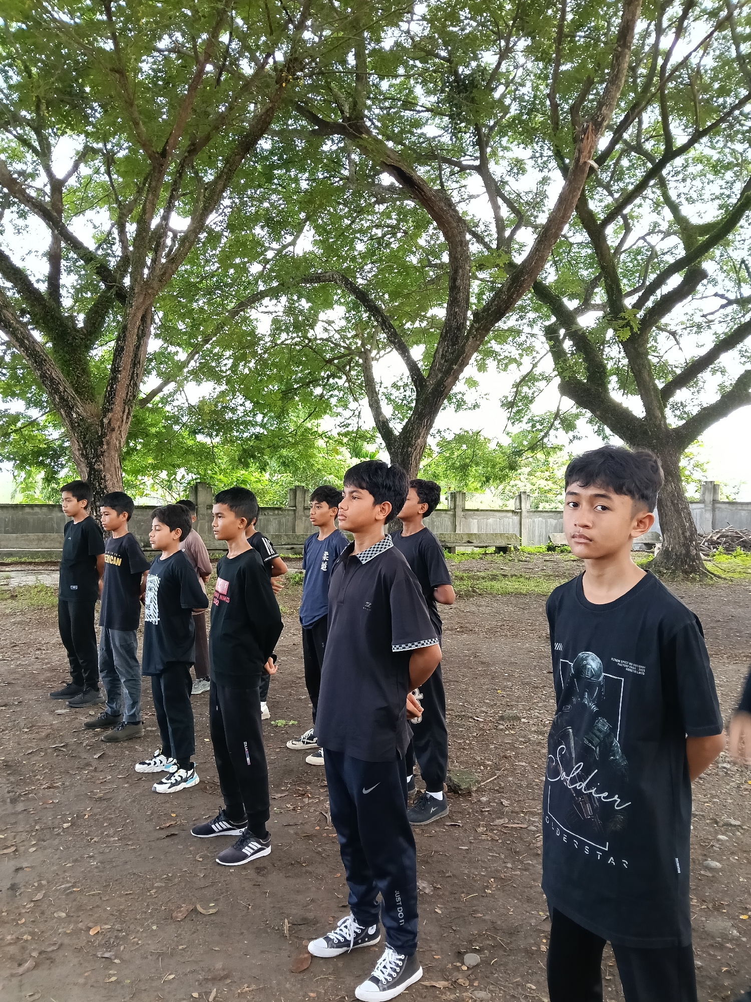 A group of eight boys are standing in a line, outdoors, in Lhokseumawe, Indonesia. They are dressed in a variety of dark-colored shirts and pants, and casual footwear. The boys are facing forward, with their hands clasped behind their backs. The ground is dirt, with fallen leaves scattered across it. Behind the boys are several large trees with spreading branches and green leaves, as well as a low concrete fence in the background. The sky is overcast.