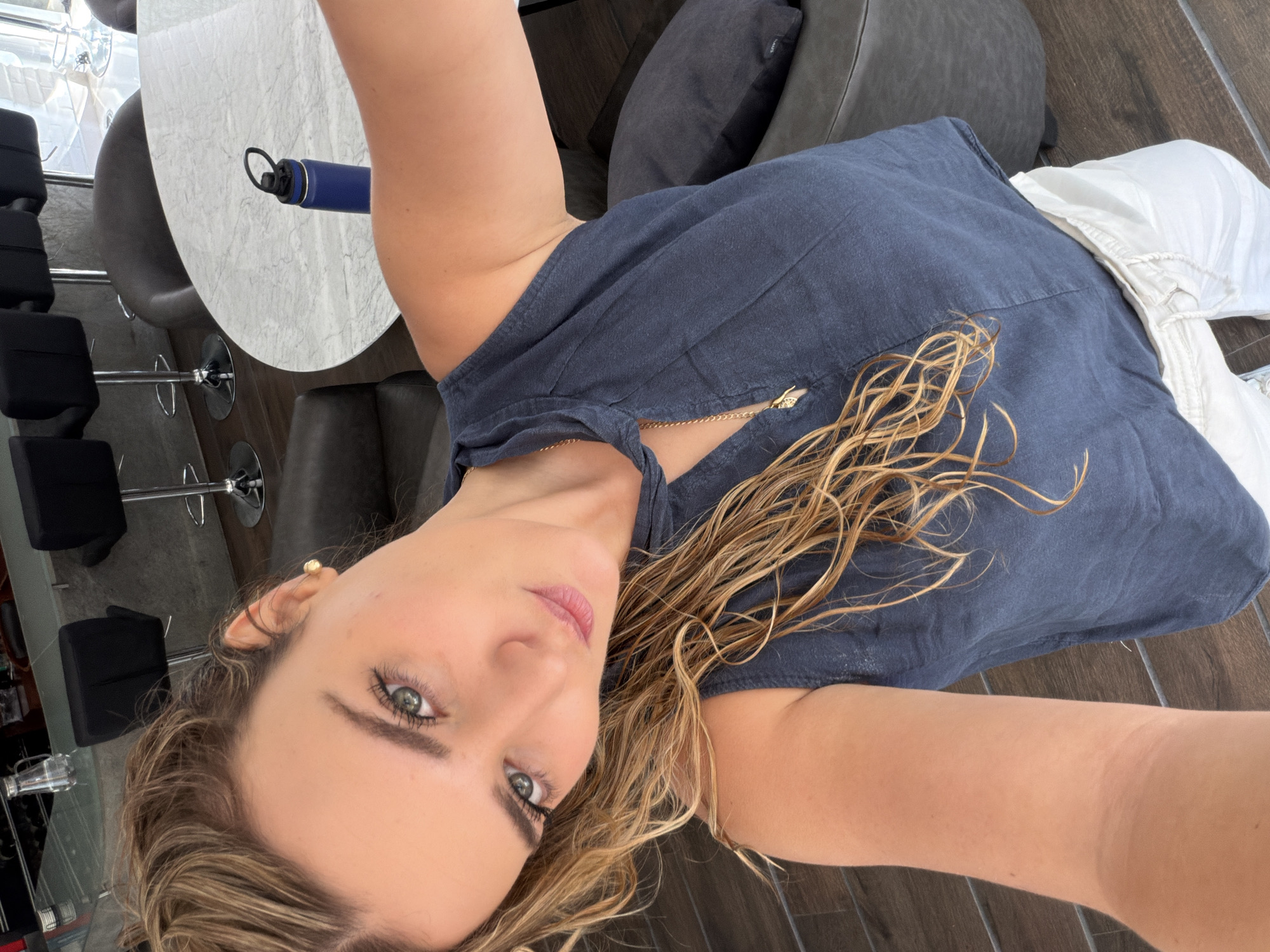 The image depicts a female individual taking a self-portrait indoors in what appears to be a modern residential or commercial space in San Bernardino Tlaxcalancingo, Mexico. The subject is a light-skinned woman with long, wavy, wet-looking light brown and blonde hair, styled to her left. Her head is tilted, and she looks directly at the camera with green/hazel eyes and light pink lips. She wears a dark blue sleeveless top with a tie-front neckline, a gold-colored chain necklace with a small pendant, a small gold stud earring in her right earlobe, and a gold ear cuff on the upper helix of her right ear. White or off-white loose-fitting pants are partially visible at the bottom of the frame.
The background shows a contemporary interior with dark brown wooden or wood-look tile flooring. To the left and right, dark gray or black upholstered seating, possibly armchairs or sections of a sofa, are visible. A dark gray round ottoman is to the subject's right. Behind her, a round table with a light gray marbled surface holds a dark blue water bottle with a black lid. Further in the background, a bar area is visible with at least four black upholstered bar stools featuring chrome bases, positioned in front of a dark gray wall. A clear glass pitcher rests on the bar surface. Bright ambient light enters from the right side of the frame, possibly from a window or glass door.