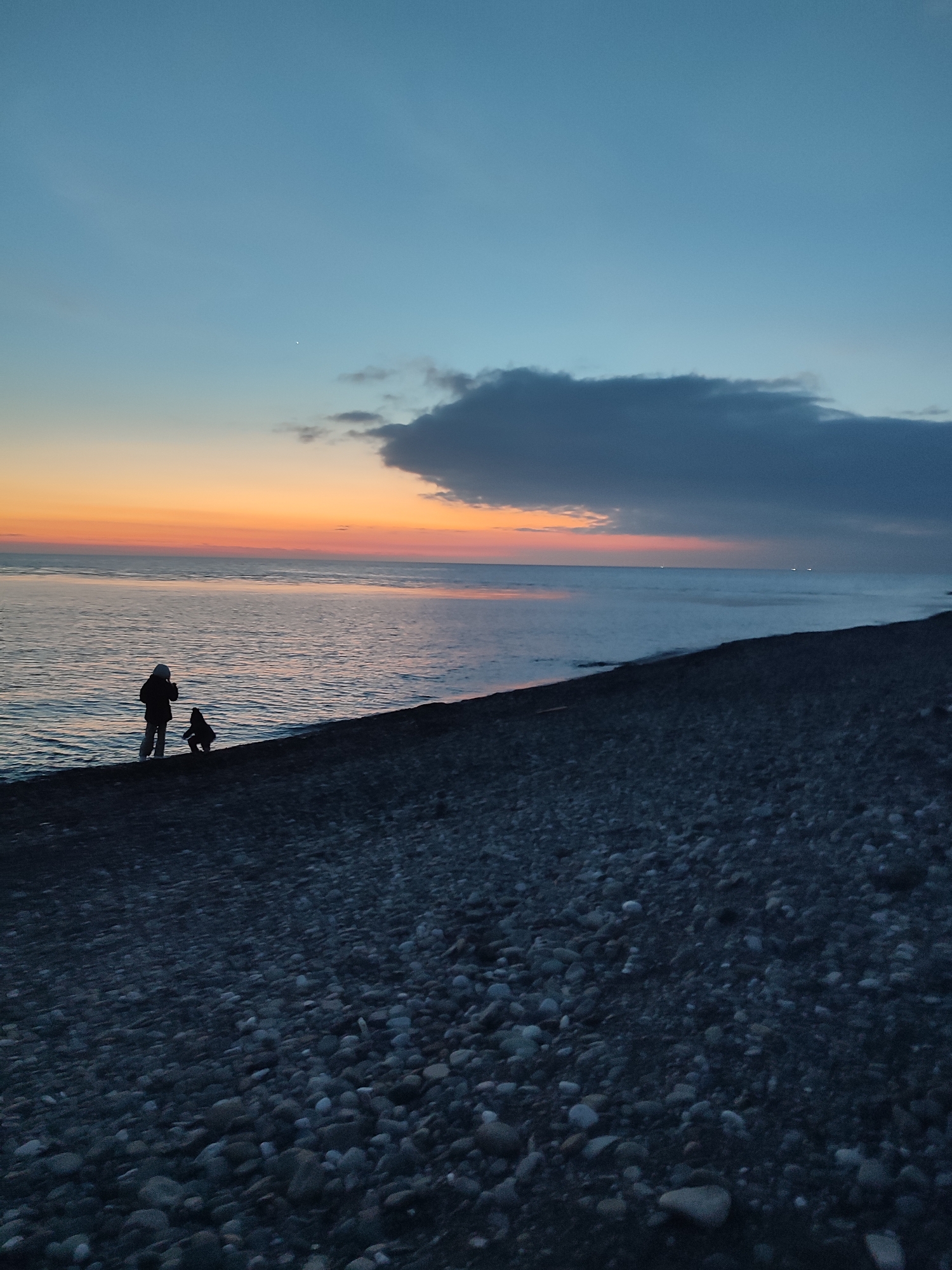 The image depicts a pebble beach at dusk in Sochi, Russia. Two figures, possibly an adult and child, stand at the edge of the water. The sky displays a gradient of color from orange near the horizon to blue above. A dark cloud hangs in the sky above the water. The beach consists of smooth, dark pebbles.