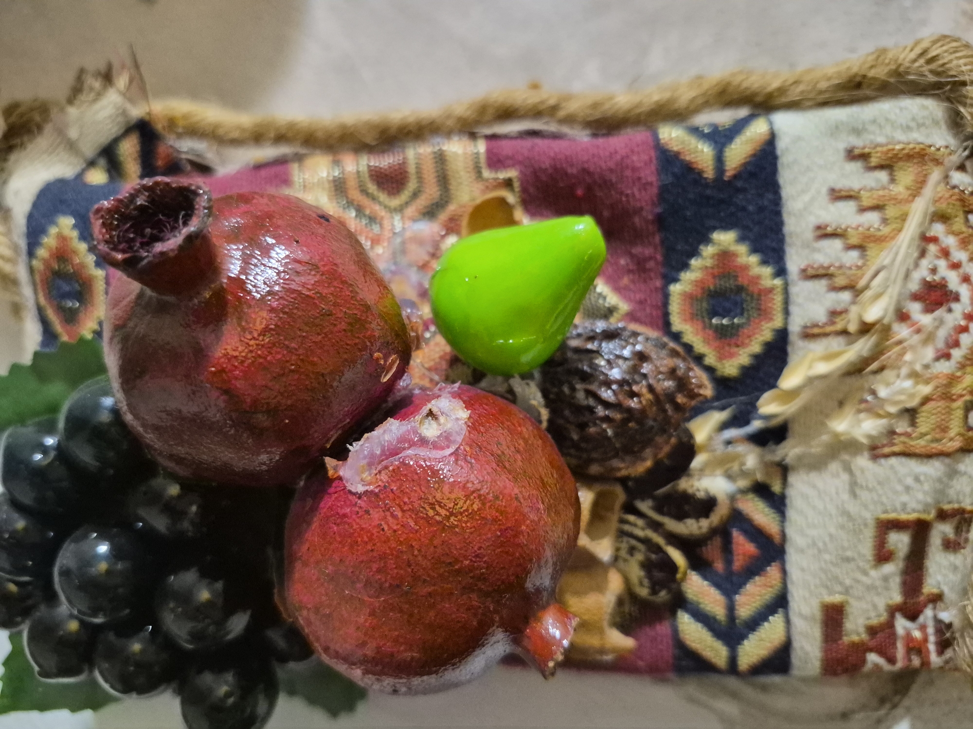 The image shows a close-up, top-down view of a decorative arrangement of fruit and what appears to be a decorative textile. The arrangement features two pomegranates, one significantly larger than the other, with bright red skin. One pomegranate has a section of its rind peeled back, revealing lighter-colored flesh. A cluster of dark, almost black, grapes is positioned above and to the left of the pomegranates, with a single green, teardrop-shaped object placed to the right of the smaller pomegranate. The objects are resting on a richly patterned textile that displays geometric designs in shades of red, blue, yellow, and cream. A textured, braided cord borders the textile on the right side. Some dried organic material, possibly straw or wheat stalks, is scattered across the textile. The background is out of focus and appears to be a light-colored surface. There is no visible indication of the city of Vanadzor, Armenia, within the image itself.