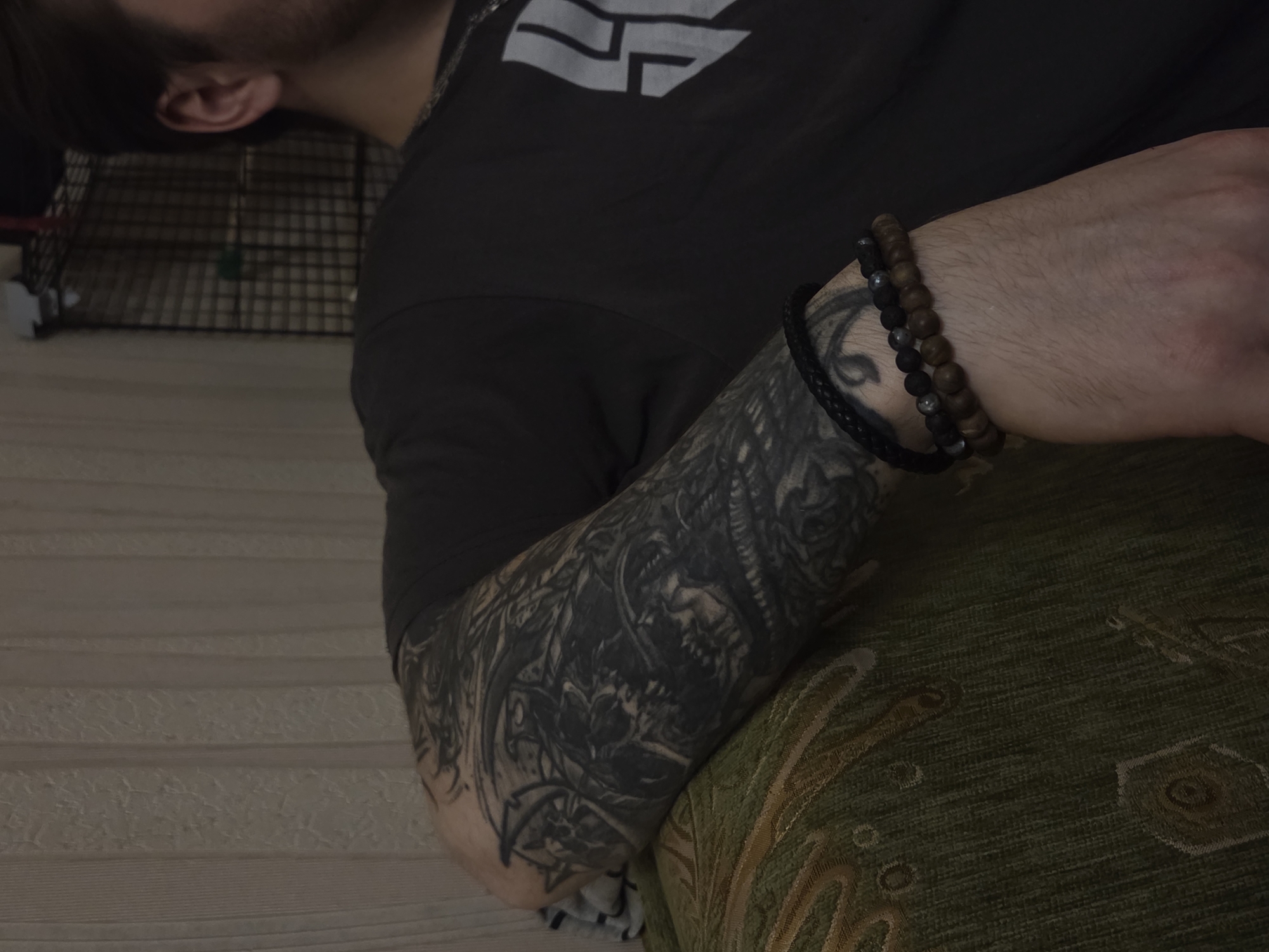 A person with a tattooed left arm rests their arm on a patterned, green-colored fabric-covered cushion or piece of furniture. The tattoo is black and grey and appears to be intricate, possibly including a skull motif. The person wears a dark gray or black shirt with a gray logo on the upper chest area. They also have a set of three bracelets on their left wrist. The background shows a section of a beige wall with linear texture and part of a black wire cage, possibly a pet enclosure, situated on a lighter-colored floor. The location is Unknown, Unknown.