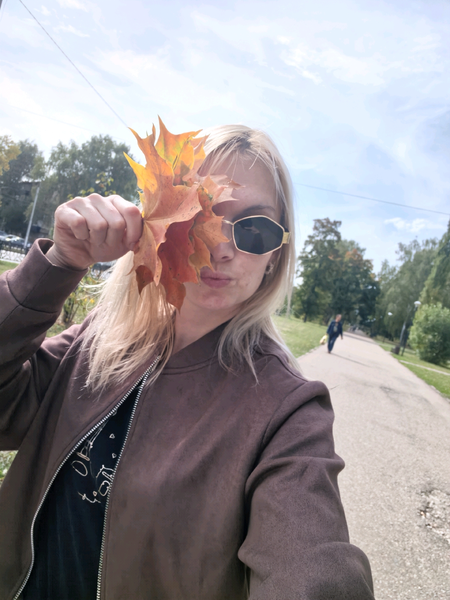 A woman with blonde hair and sunglasses holds a cluster of orange and yellow maple leaves in front of her face, obscuring her eyes. She is wearing a brown bomber jacket over a black t-shirt with white writing. The woman is standing on a paved path in a park, with trees and a bright sky visible in the background. Another person is walking away from the camera on the path in the distance. The image appears to be a selfie, taken in Sterlitamak, Russia, likely during the autumn season given the fall foliage.