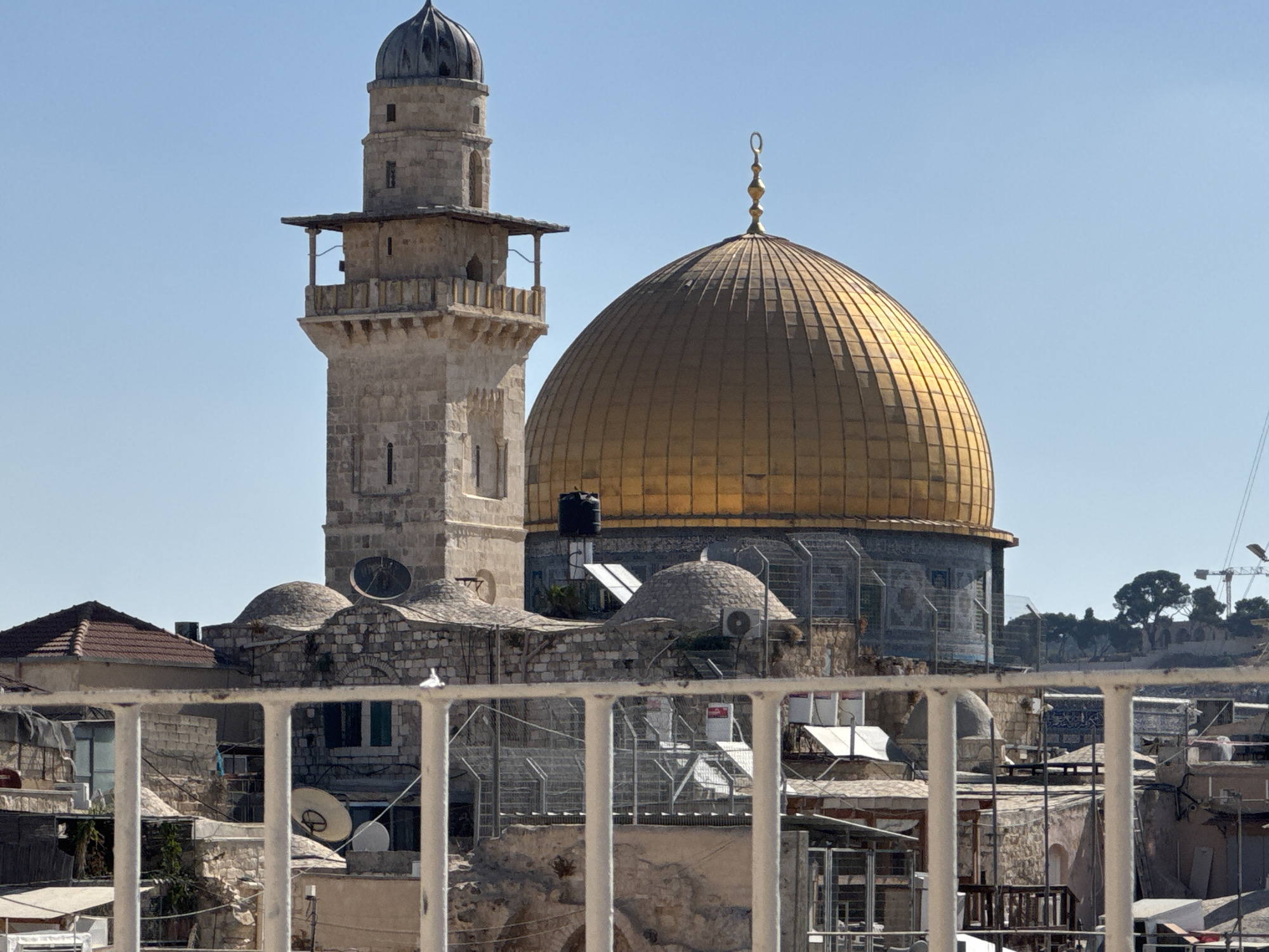 The image shows a view of the Dome of the Rock in Jerusalem, Israel, a prominent landmark on the Temple Mount. The golden dome is a dominant feature, set against a clear blue sky. To the left of the dome, a tall, stone minaret with a balcony and a smaller dome on top is visible. The foreground is partially obscured by a white railing, behind which lie rooftops of various buildings made of stone and tile. Some of these buildings appear to be residential or commercial, with satellite dishes and air conditioning units visible. The overall scene suggests an urban environment, possibly from a higher vantage point overlooking the city.