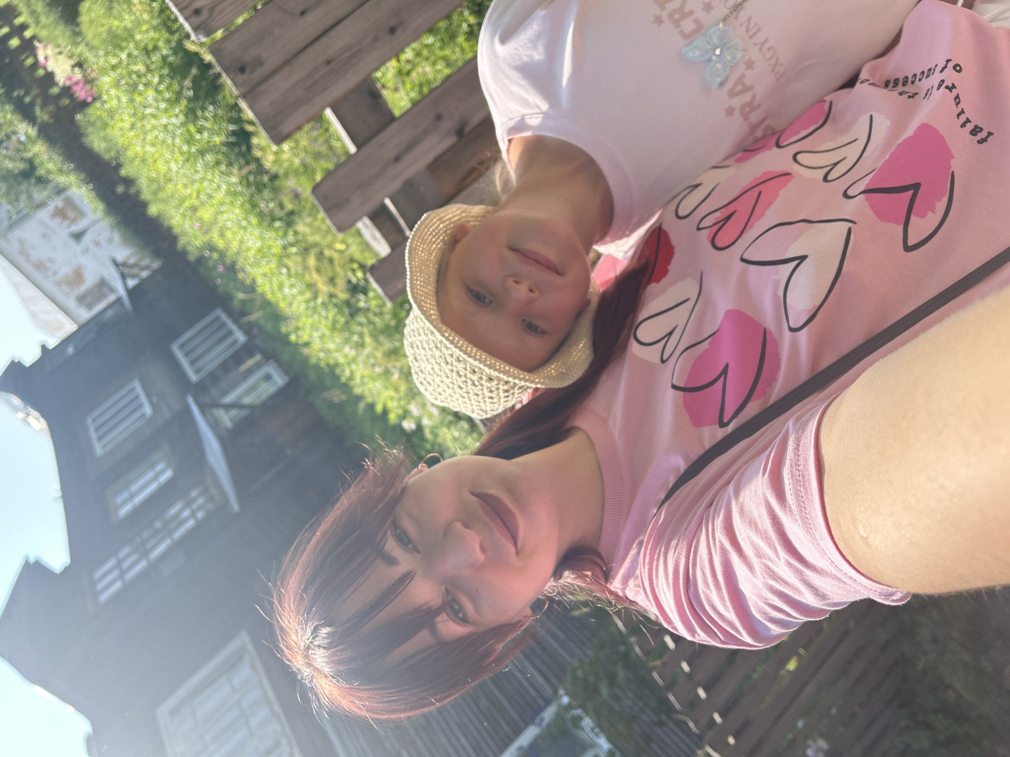 In the city of Krasnovishersk, Russia, a selfie is taken outdoors on a sunny day. Two females are pictured. The foremost subject, a woman with auburn hair, smiles towards the camera. She is wearing a pink t-shirt with a heart design. To her right, a young girl with blonde hair, wearing a straw hat and a light pink t-shirt, leans her head on the woman's shoulder. In the background, a rustic wooden fence runs along the right side of the image, and a weathered, dark wooden house with visible windows stands in the left background, partially obscured by a bright sun glare. Lush green foliage surrounds the house and extends into the background. The overall scene depicts a casual, outdoor gathering in a rural or semi-rural setting.
