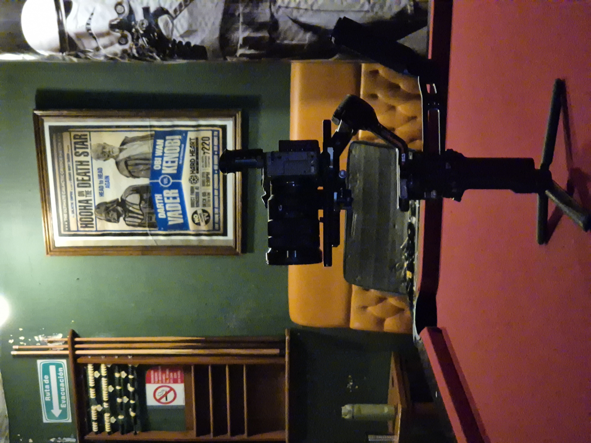 The image depicts a room with various objects. A black camera rig is positioned prominently in the foreground. In the background, a framed poster parodying a Star Wars theme hangs on a green wall. To the left, there is a wooden rack holding pool cues and a sign labeled "Ruta de Evacuación" (Evacuation Route) in Spanish, along with a "no smoking" symbol. Above the framed poster is a partially visible decorative display featuring what appears to be a Stormtrooper helmet and other memorabilia. The location is suggested to be Puebla, Mexico. Other objects include a red surface, possibly a pool table, and a yellow cushioned object.