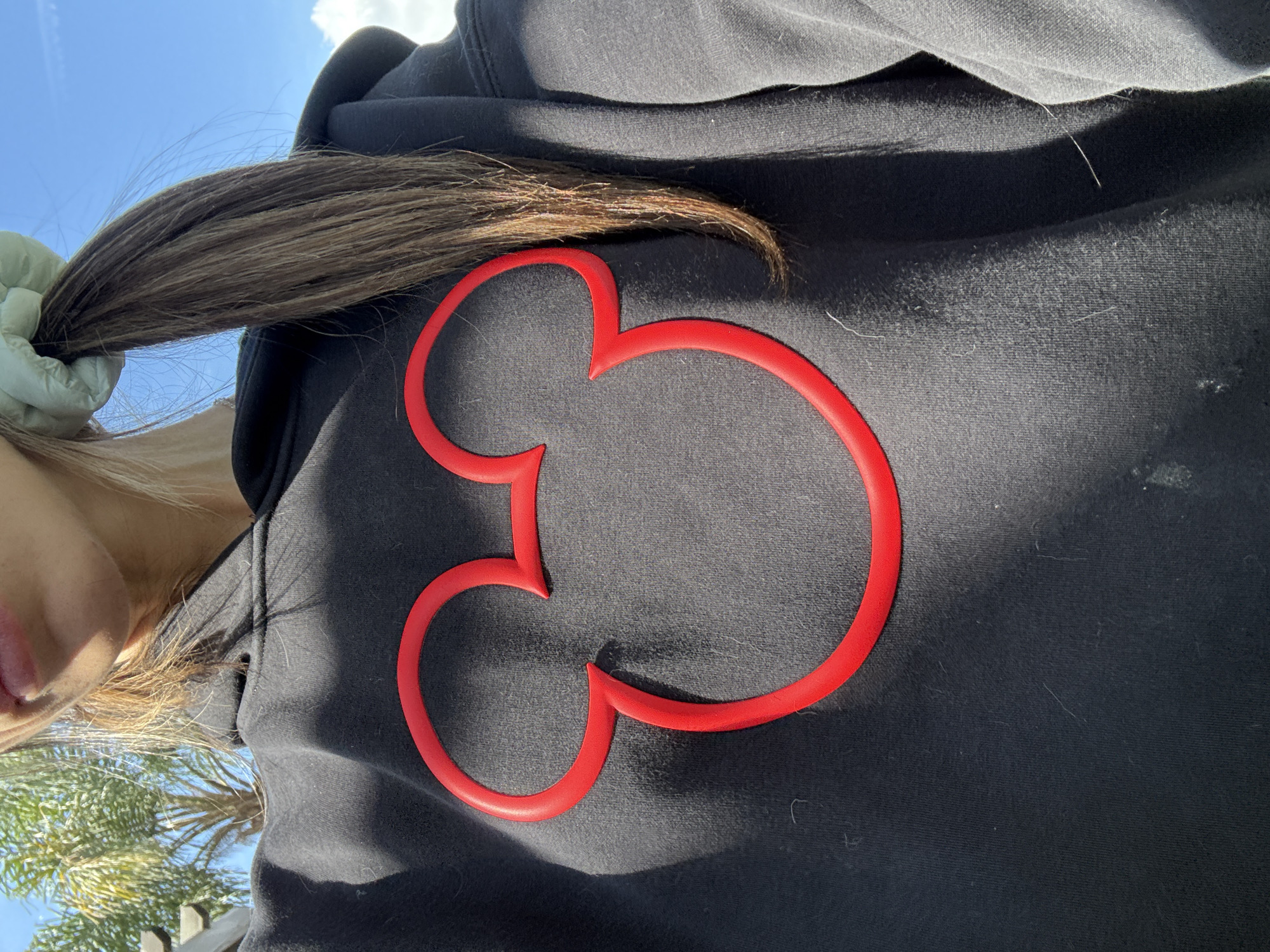 This image captures a close-up, outdoor shot of a person wearing a black hoodie, featuring a prominent red Mickey Mouse silhouette outline. The person is seen from a high angle, with their head partially visible on the left side of the frame, looking slightly upwards. Long, dark brown hair, gathered and tied back with a light green scrunchie, extends across the top of the hoodie.
The central element is the red Mickey Mouse outline, which appears to be a raised, three-dimensional appliqué or object attached to the black fabric, casting subtle shadows on the hoodie, emphasizing its texture. The black hoodie itself shows some visible lint and fibers on its surface.
The setting is outdoors under bright, direct sunlight, suggesting midday or early afternoon. The upper left corner of the image reveals a clear blue sky with some scattered white clouds. In the blurred background on the lower left, distinct palm trees are visible, providing a strong cue of a warm, possibly tropical or Californian location, consistent with Seal Beach, United States of America.
No specific activity is clearly taking place beyond the subject posing for the photo. The overall mood appears casual and relaxed. There is no visible text in the image.