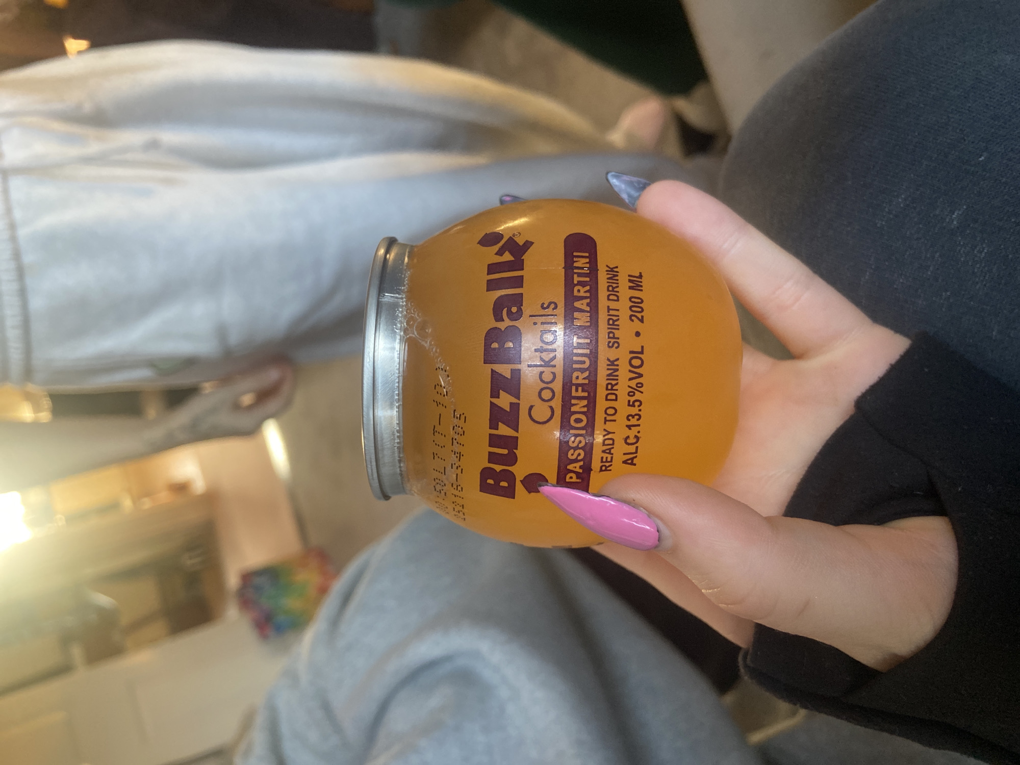 A hand is holding a "BuzzBallz Cocktails" Passionfruit Martini can. The hand has one fingernail painted pink and one with a dark blue color. The can reads "READY TO DRINK SPIRIT DRINK" and "ALC.13.5%VOL. 200 ML." The person holding the can is sitting down and has a black sleeve of clothing, while wearing grey trousers. Background features are out of focus, but show the suggestion of the interior of a building and other people. It's impossible to determine the location in the city of Reigate and Banstead, United Kingdom based on the details provided in the image.