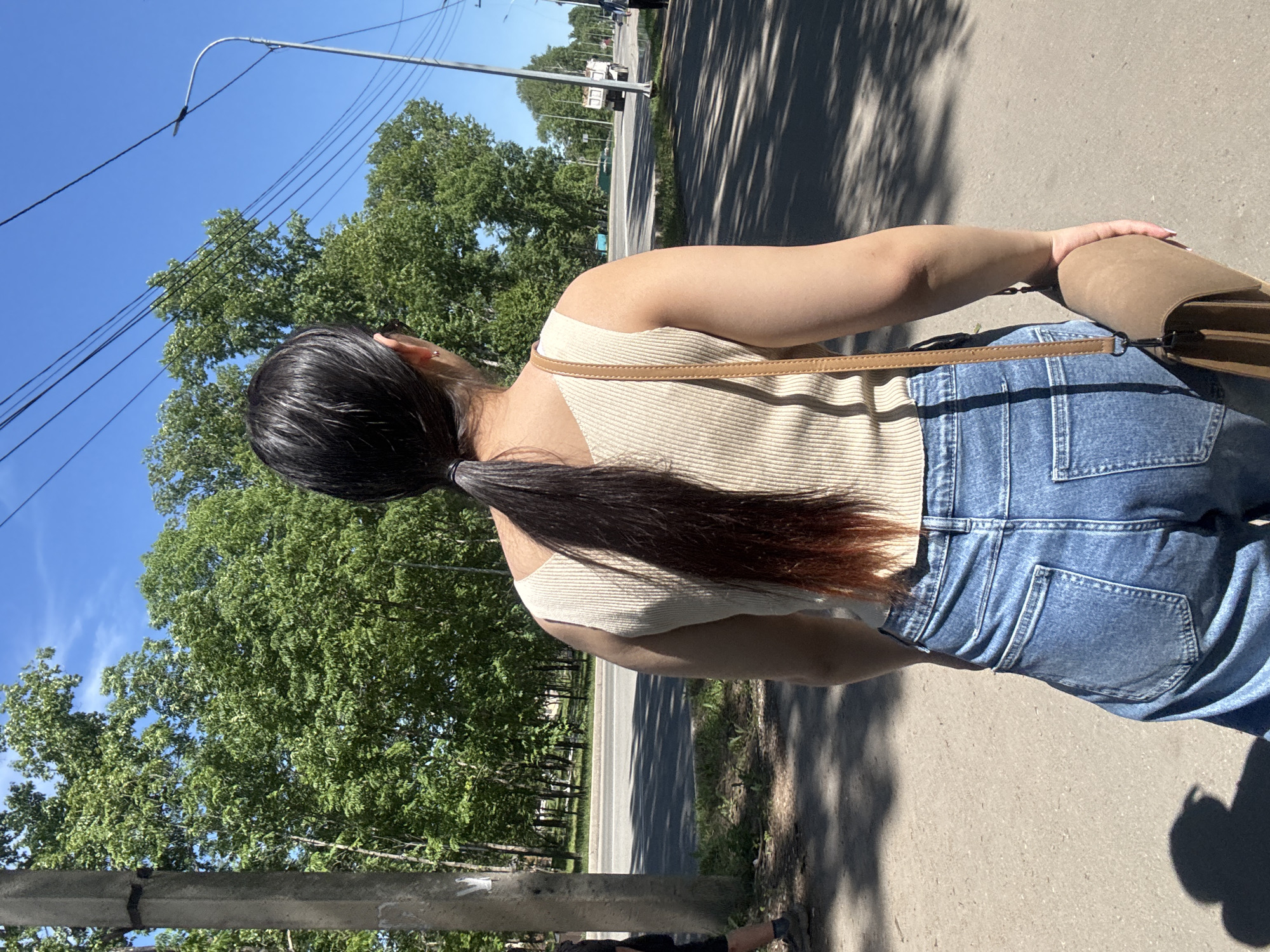 In Birobidzhan, Russia, a young woman with dark hair in a ponytail walks away from the camera on a paved path. She wears a cream-colored ribbed tank top and loose-fitting blue jeans. A tan crossbody bag is slung over her right shoulder. Behind her, a two-lane road runs parallel to a line of lush green trees. The sky is clear blue, and a street light stands on the right side of the road. Sunlight casts shadows on the path.