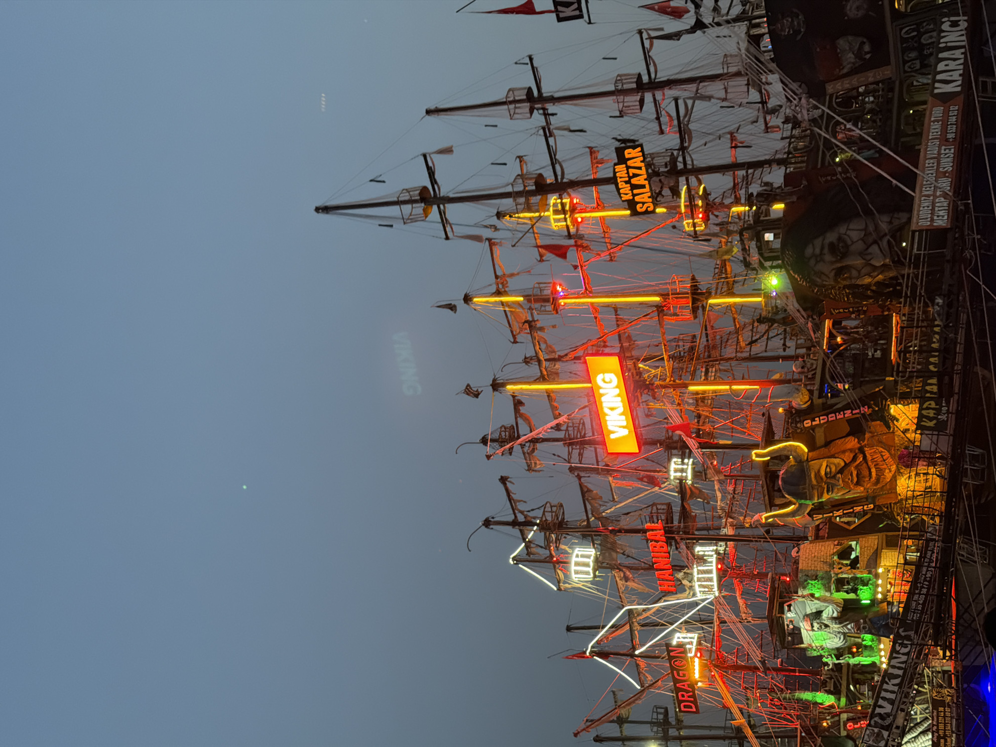 This image captures a twilight scene of elaborately decorated pirate-themed boats docked in the city of Fethiye, Turkey. The boats are adorned with bright neon lights in red, yellow, and white, illuminating their masts, rigging, and decorative elements. Prominent signage includes "VIKING," "HANIBAL," and "KAPTAN SALAZAR," suggesting these are tour or entertainment vessels. Large, stylized Viking and other character figures are visible on the decks. The sky above is a muted blue and grey, indicating the transition from day to night. The overall impression is one of a vibrant tourist attraction, likely located along Fethiye's waterfront or harbor area.